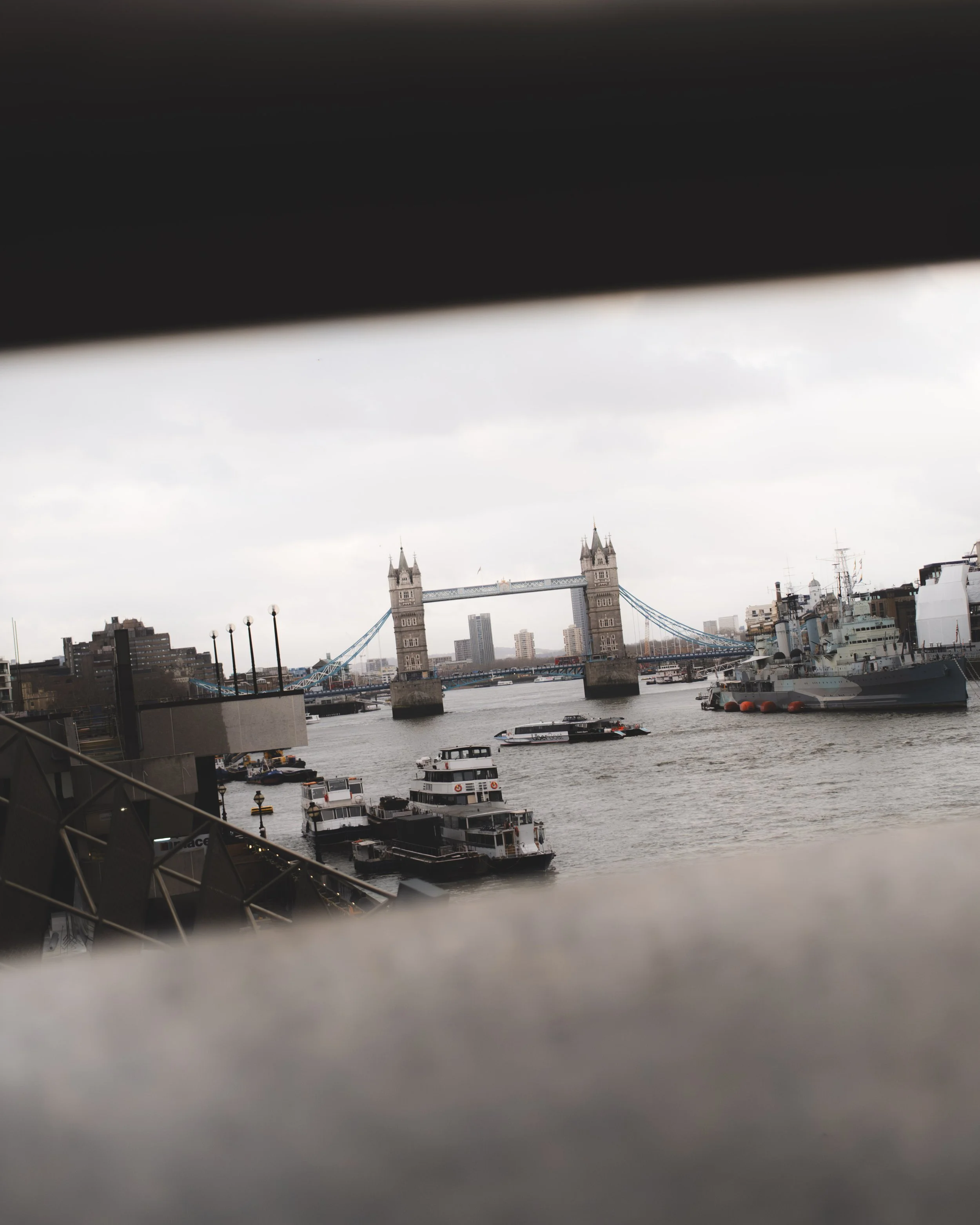 View of Tower Bridge in London with boats on the River Thames under an overcast sky, partially framed by dark objects in the foreground.