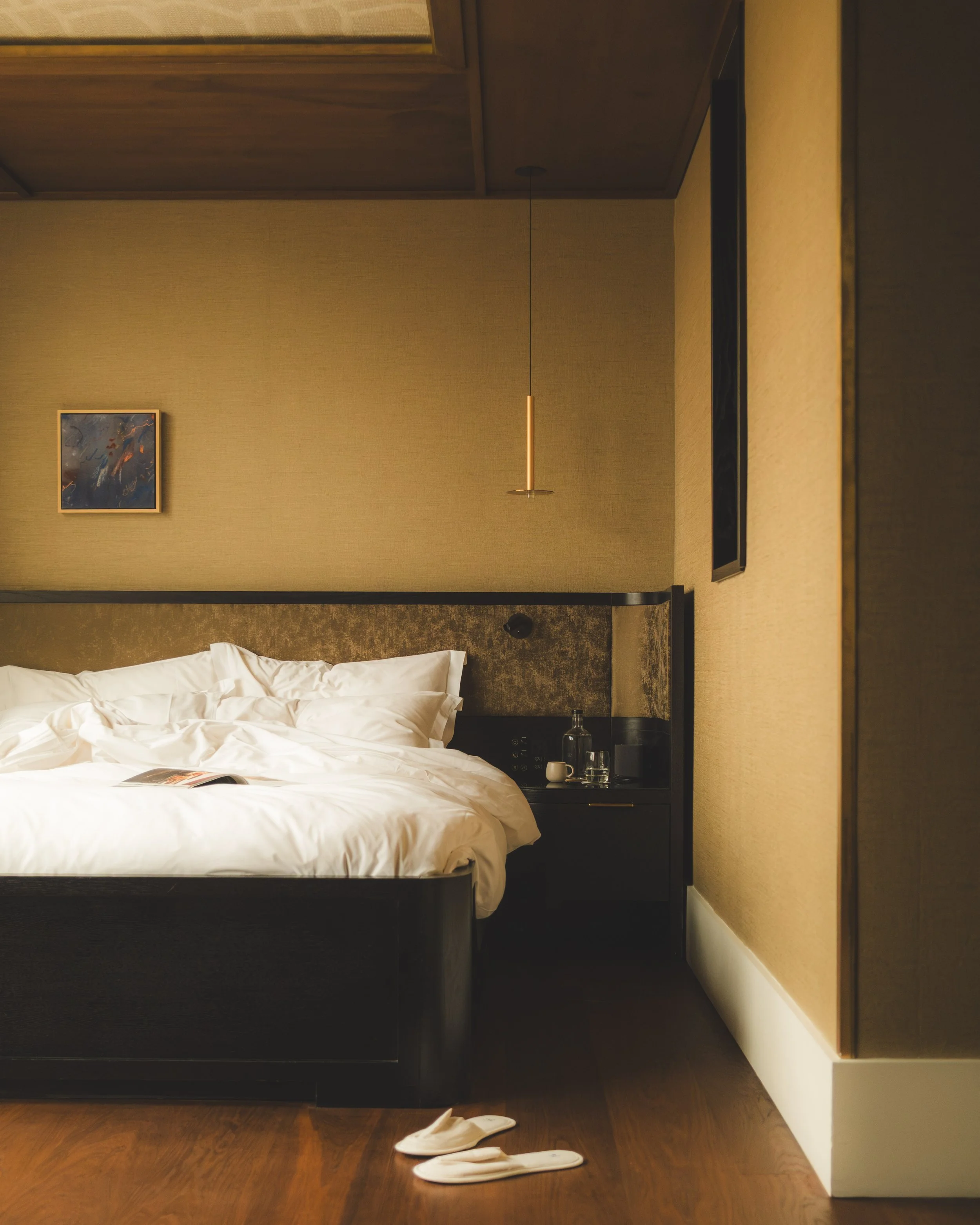 A hotel room with a bed, a nightstand with a glass carafe and two glasses, a small white cup, a long thin hanging light fixture, a framed abstract painting, and a pair of white slippers on the wooden floor.