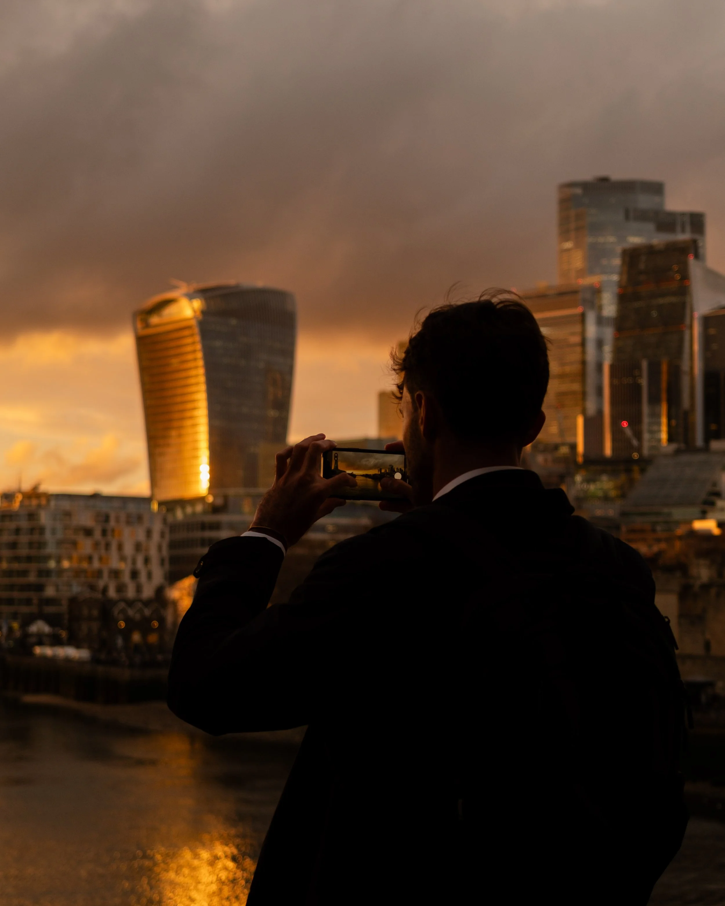 Silhouette of a man taking a photo with smartphone of modern city skyline during sunset, with dark clouds overhead. Lionel Chu - Hotel & Travel Photographer