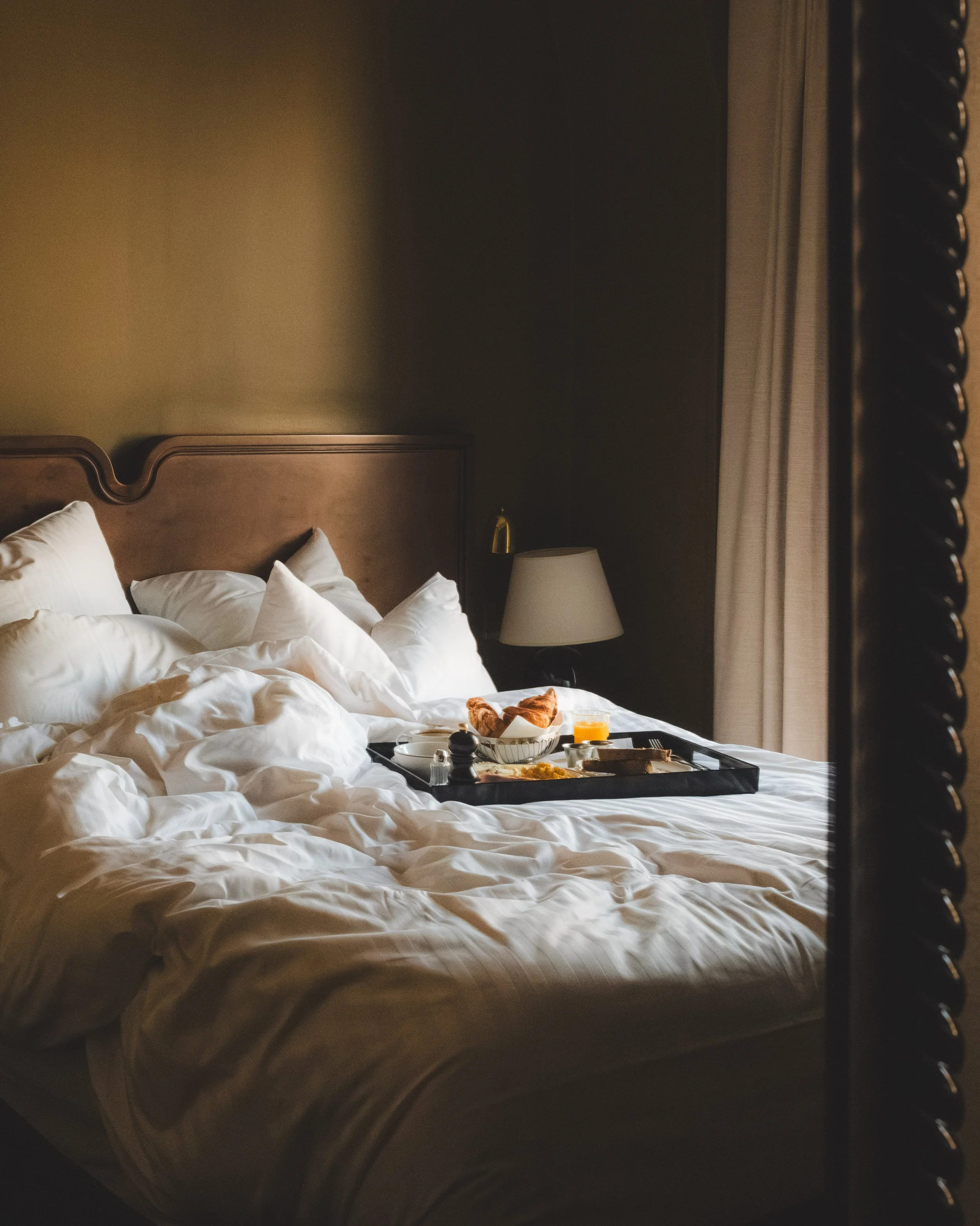 A neatly made bed with white pillows and a brown headboard, with a tray of breakfast including a croissant, orange juice, and other breakfast items on the bed. A table lamp and curtains are visible in the background.
