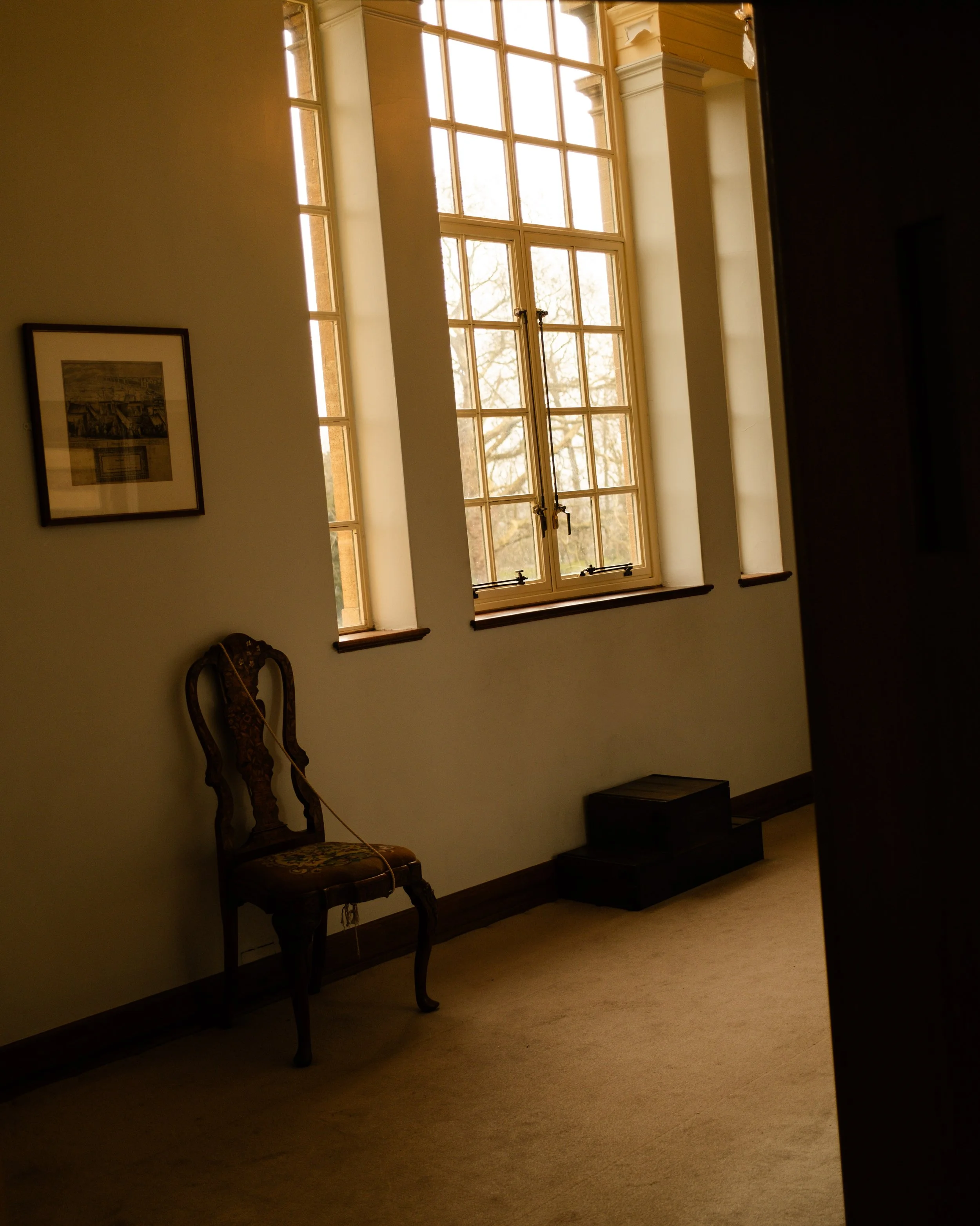 Room with large windows letting in natural light, an ornate wooden chair with a floral cushion, a framed picture on the wall, and stairs leading down. Lionel Chu - Hotel & Travel Photographer