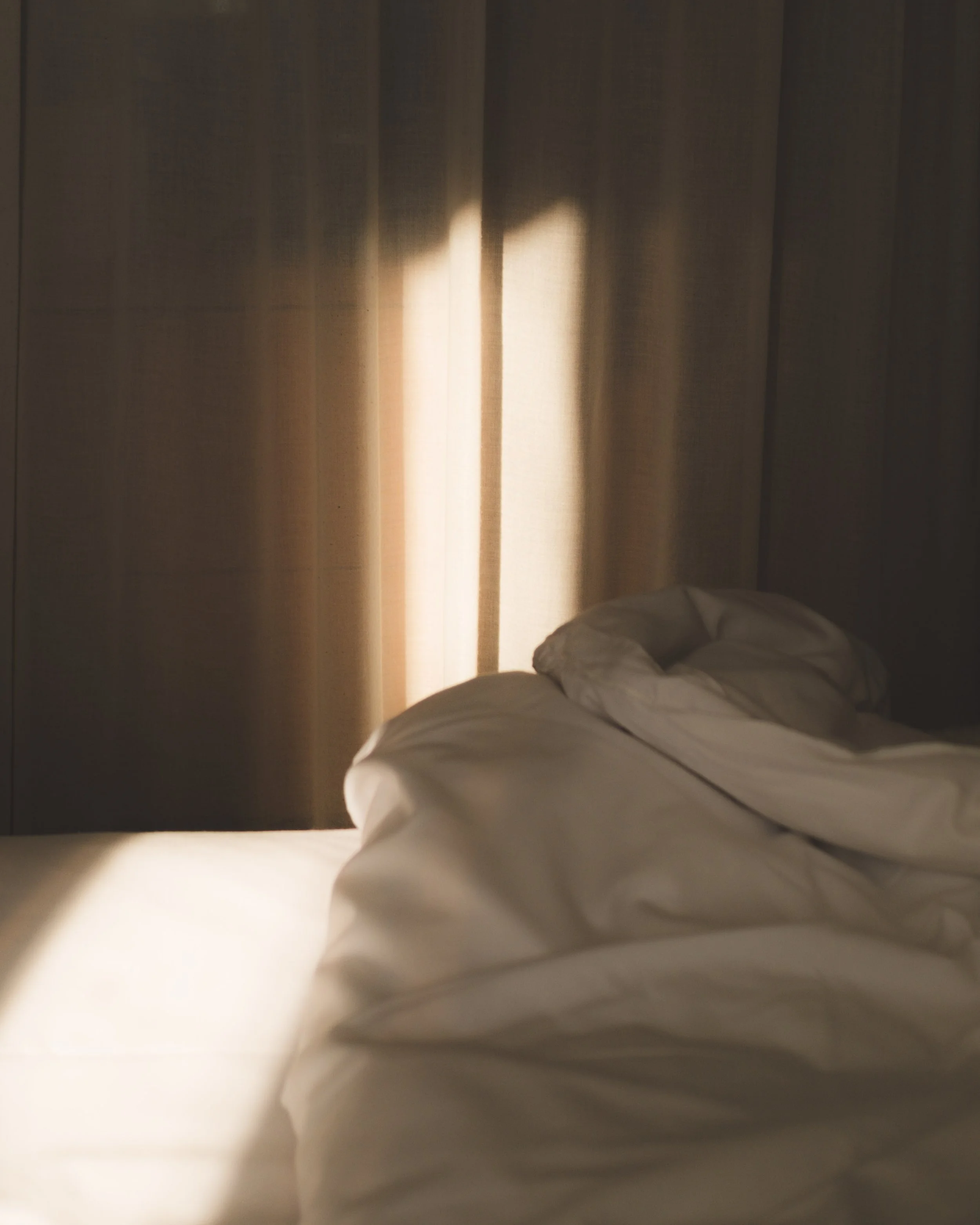A bed with white sheets and pillows is seen in front of beige curtains with sunlight filtering through.