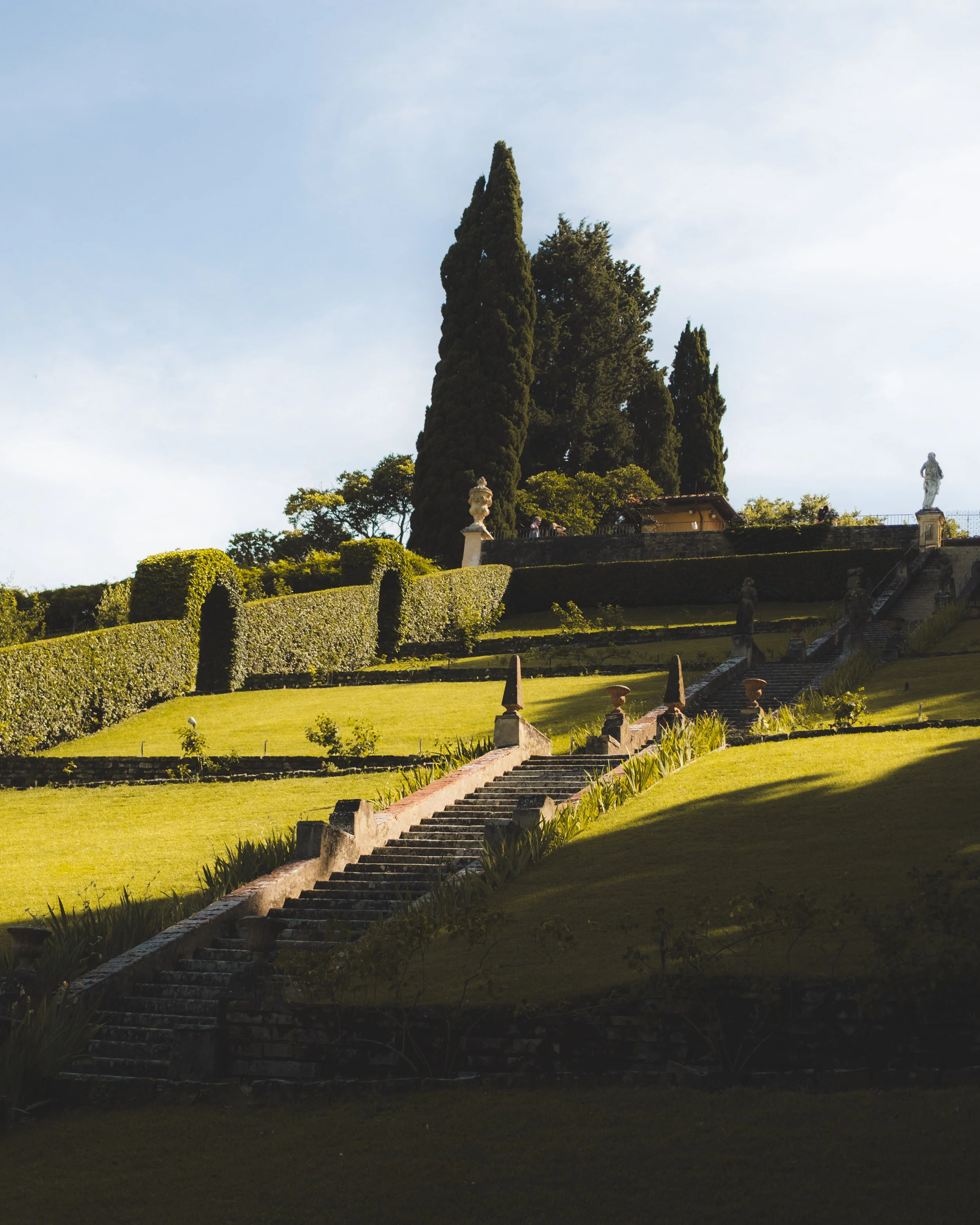 A series of stairs ascend a lush, terraced hillside garden with tall cypress trees and statues, under a cloudy sky. Lionel Chu - Hotel & Travel Photographer