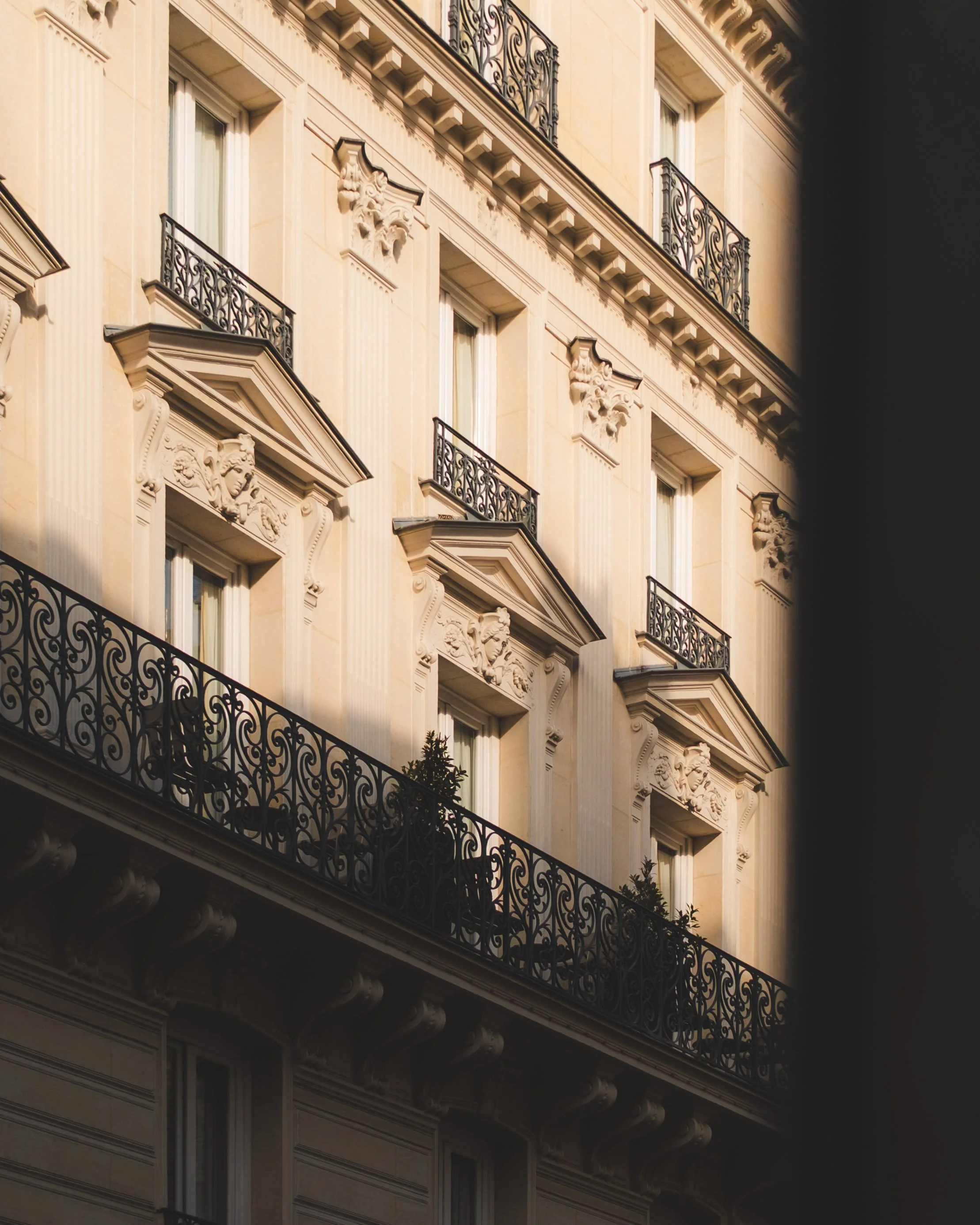 Close-up view of a historic beige building with ornate architectural details, multiple balconies with wrought iron railings, and decorative sculptures above the windows, illuminated by sunlight.