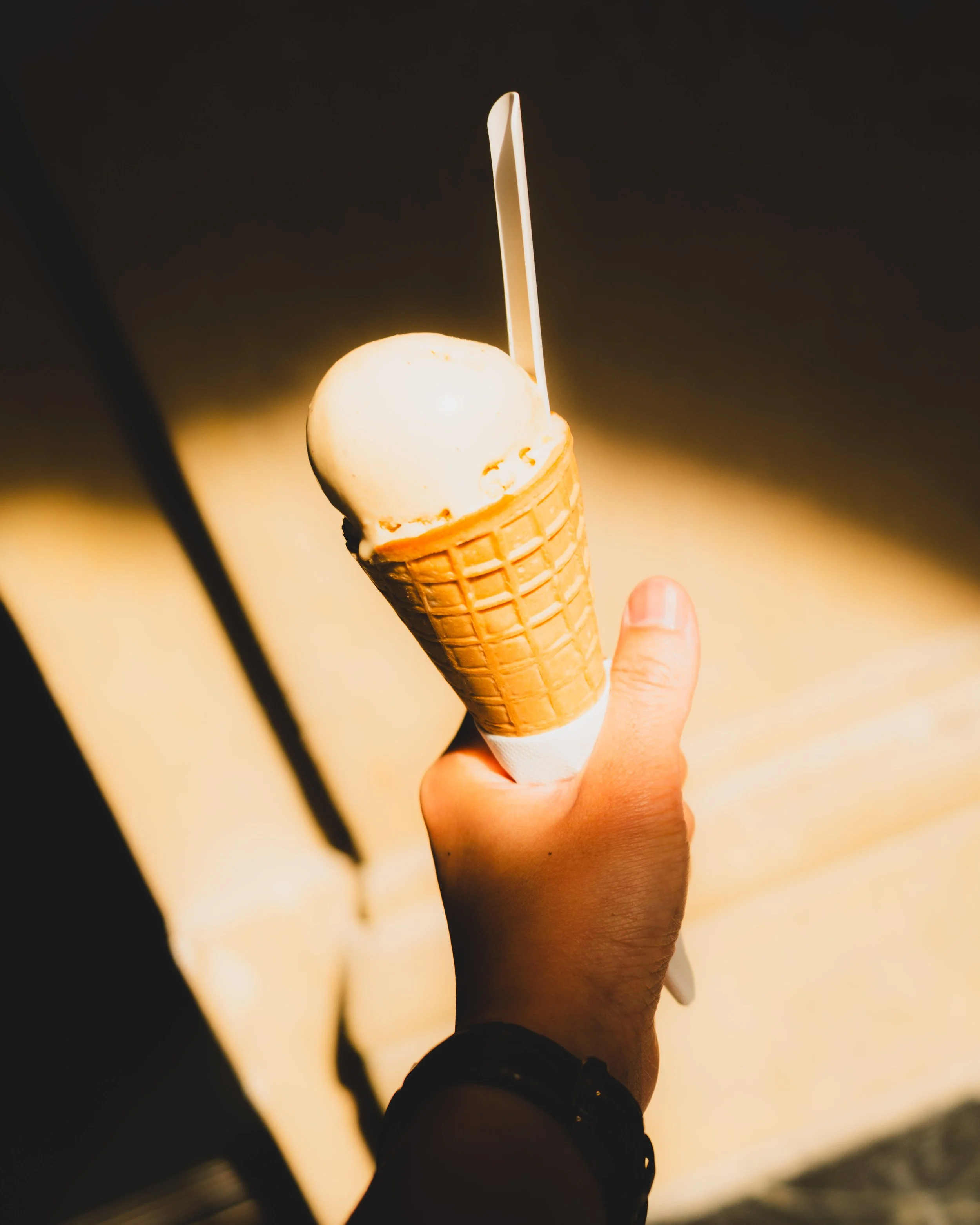 A hand holding a cone with a single scoop of vanilla ice cream, with a plastic spoon inserted, against a dark background. Lionel Chu - Hotel & Travel Photographer
