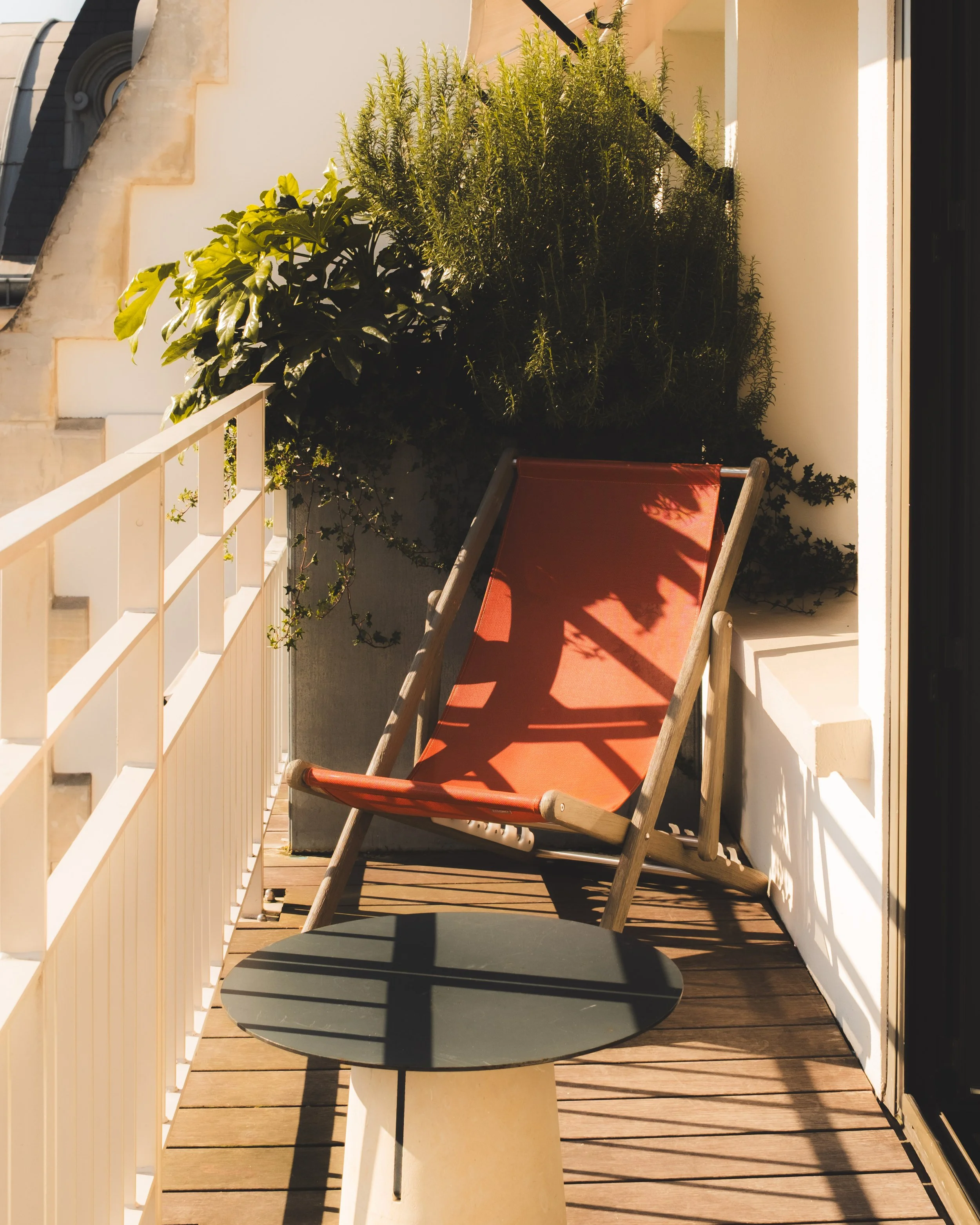 An hotel balcony with a small round table, a folding chair with a red seat, and greenery in the background, illuminated by sunlight.