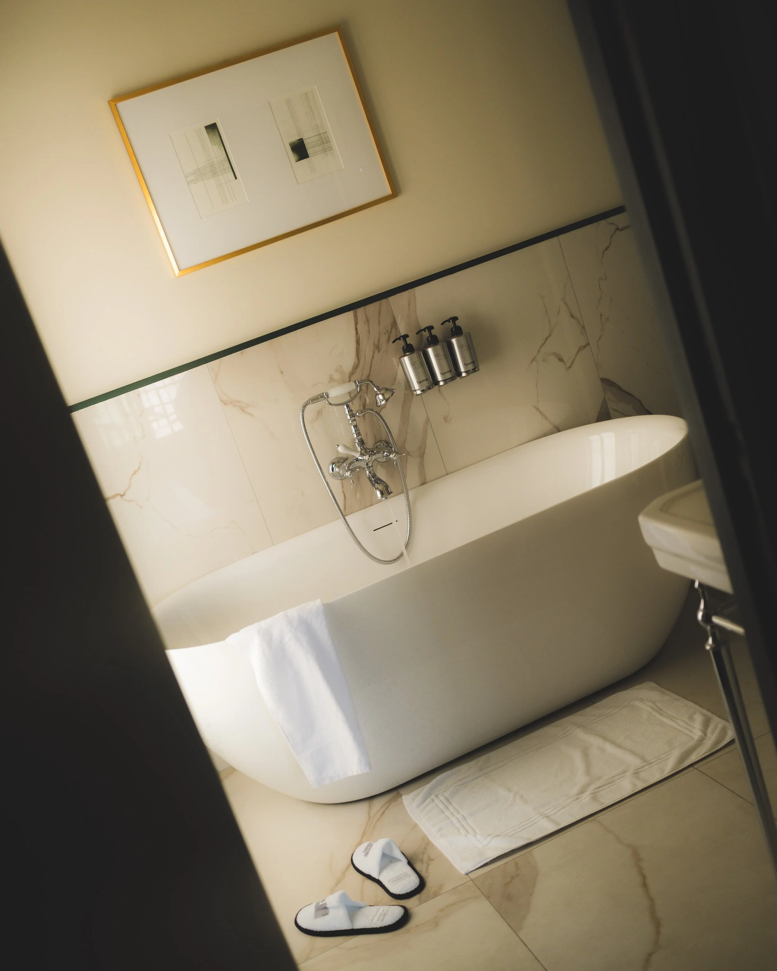 A bathroom with marble walls, a white bathtub, black-framed mirror, three soap dispensers, a white towel hanging over the tub, and two socks on the marble floor.