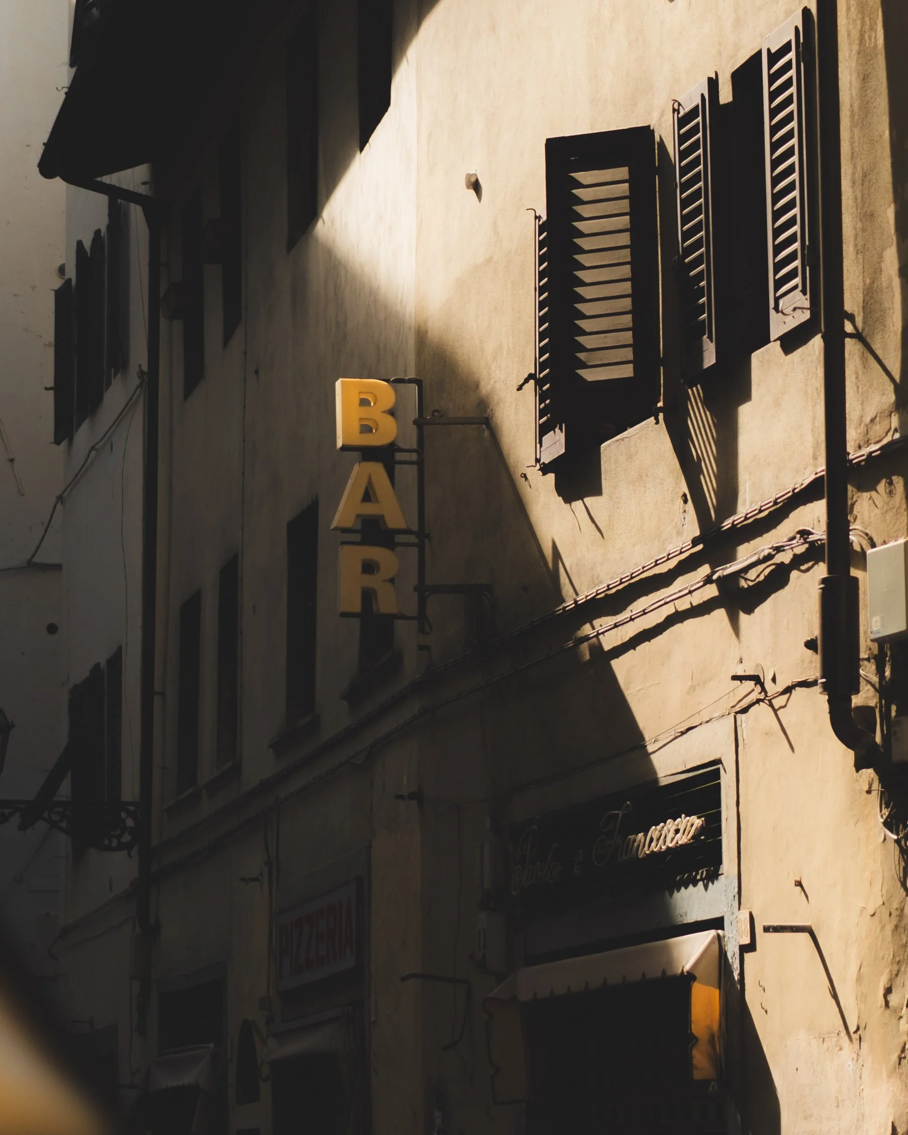 Sunlit beige building with black shutters, a vertical yellow sign spelling 'BAR', and a black sign with white cursive text. Lionel Chu - Hotel & Travel Photographer