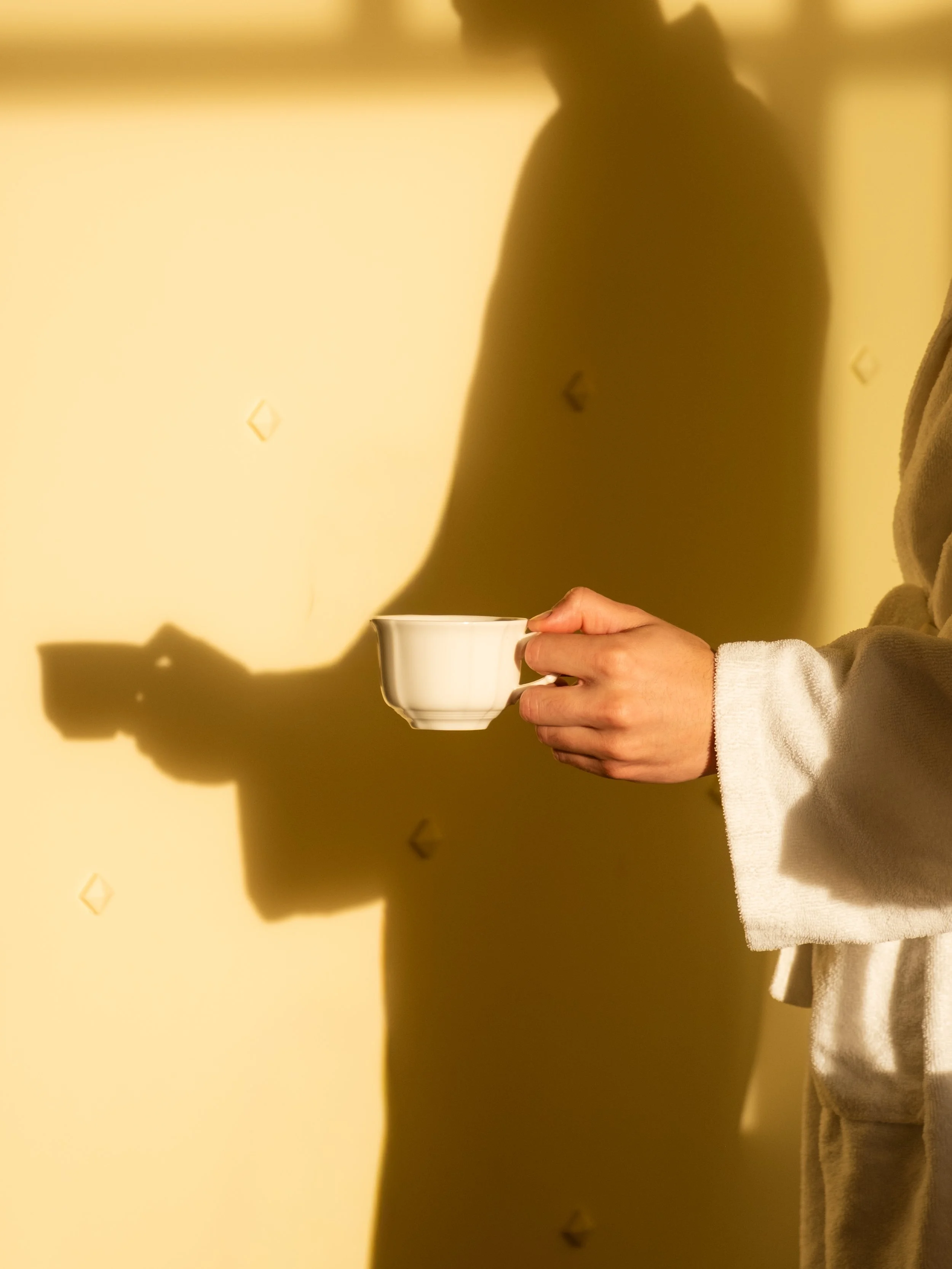 Person holding a white teacup with a shadow cast on a beige wall. Lionel Chu - Hotel & Travel Photography