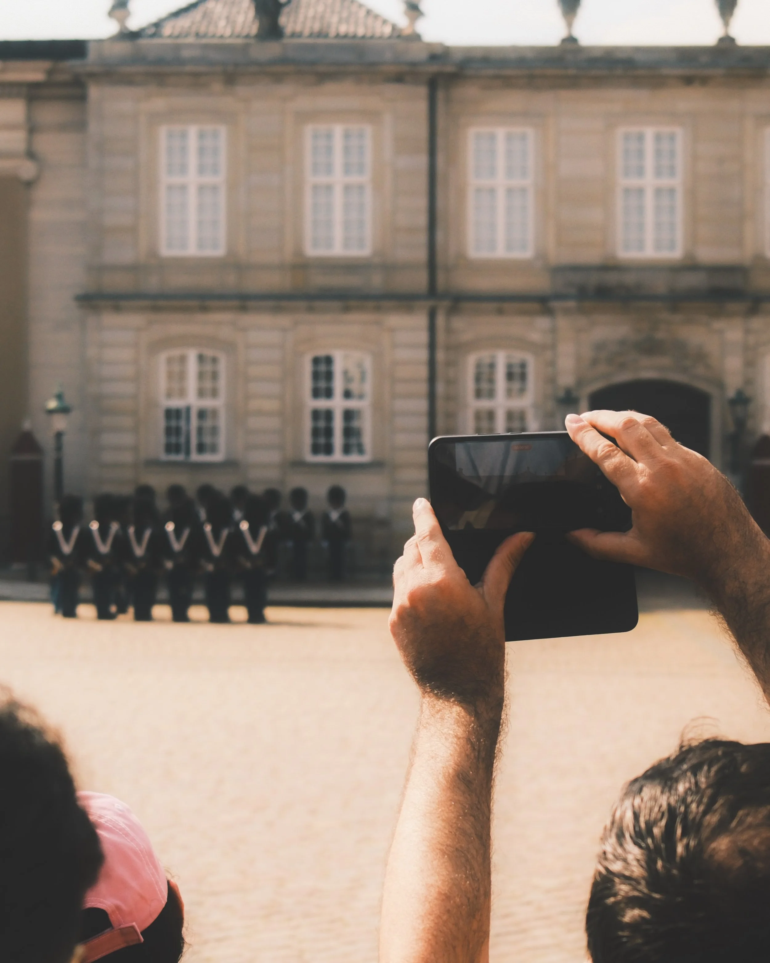 Person taking a photo with a smartphone of a historic building with multiple windows, seen from behind a crowd of people.