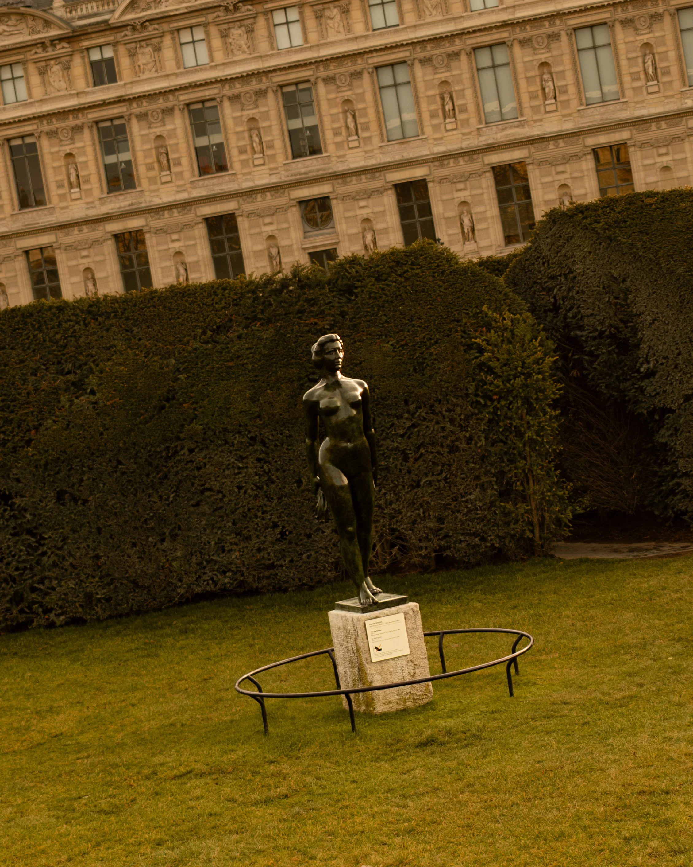 Bronze sculpture of a woman standing outdoors in the Tuileries Gardent on a stone pedestal, surrounded by a low metal railing, with a hedge and the Louvre Museum in the background. Lionel Chu - Hotel & Travel Photographer
