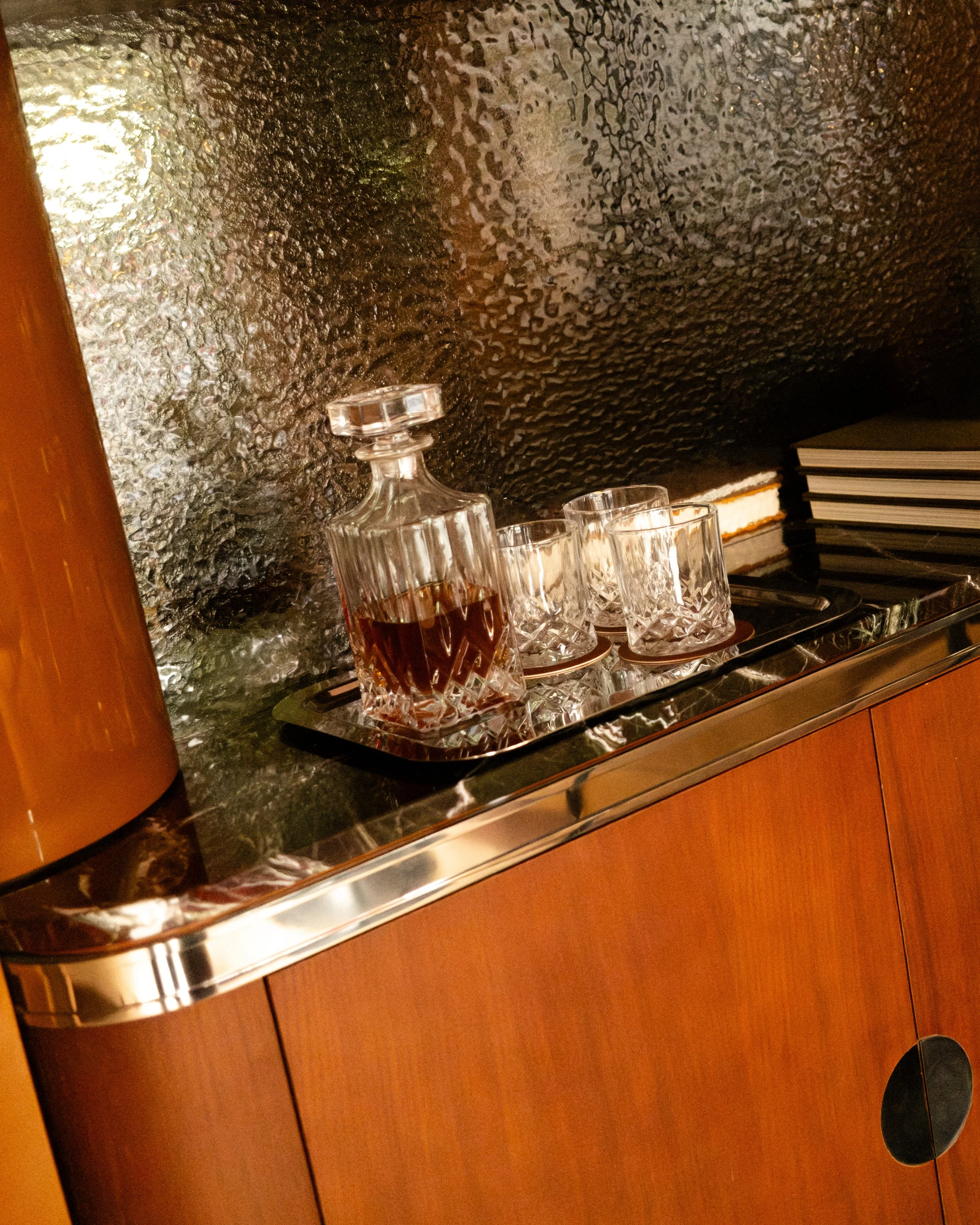 A wooden bar counter with a glass decanter and four glass tumblers placed on a tray, with a frosted glass wall and a stack of black books or notebooks in the background. Lionel Chu - Hotel & Travel Photography