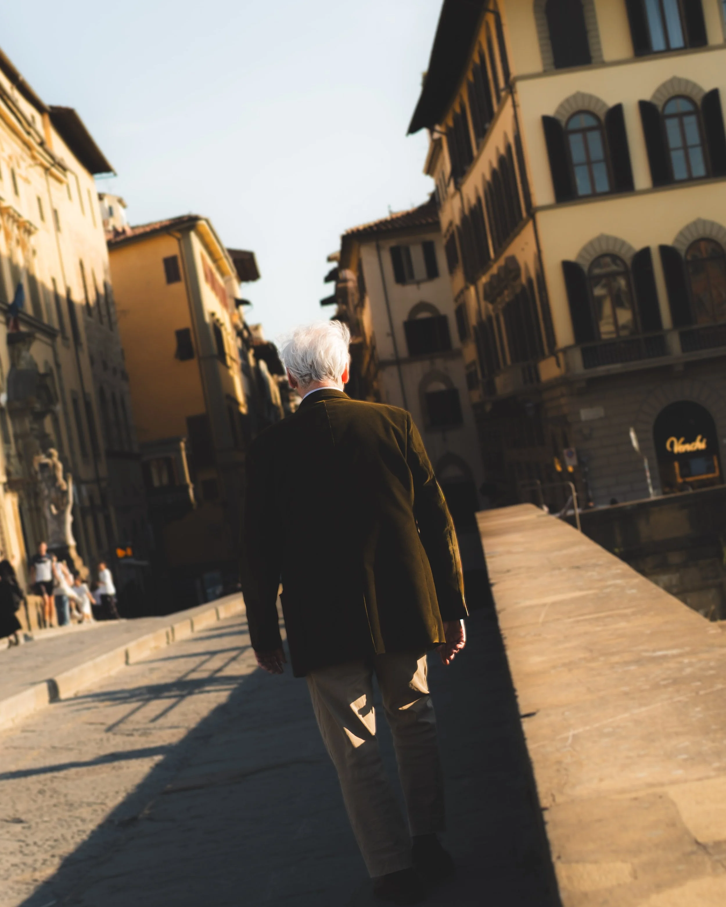 An elderly man with white hair, dressed in a dark jacket and light-colored pants, walking along a European city street during sunset, with old buildings and other pedestrians in the background. Lionel Chu - Hotel & Travel Photographer
