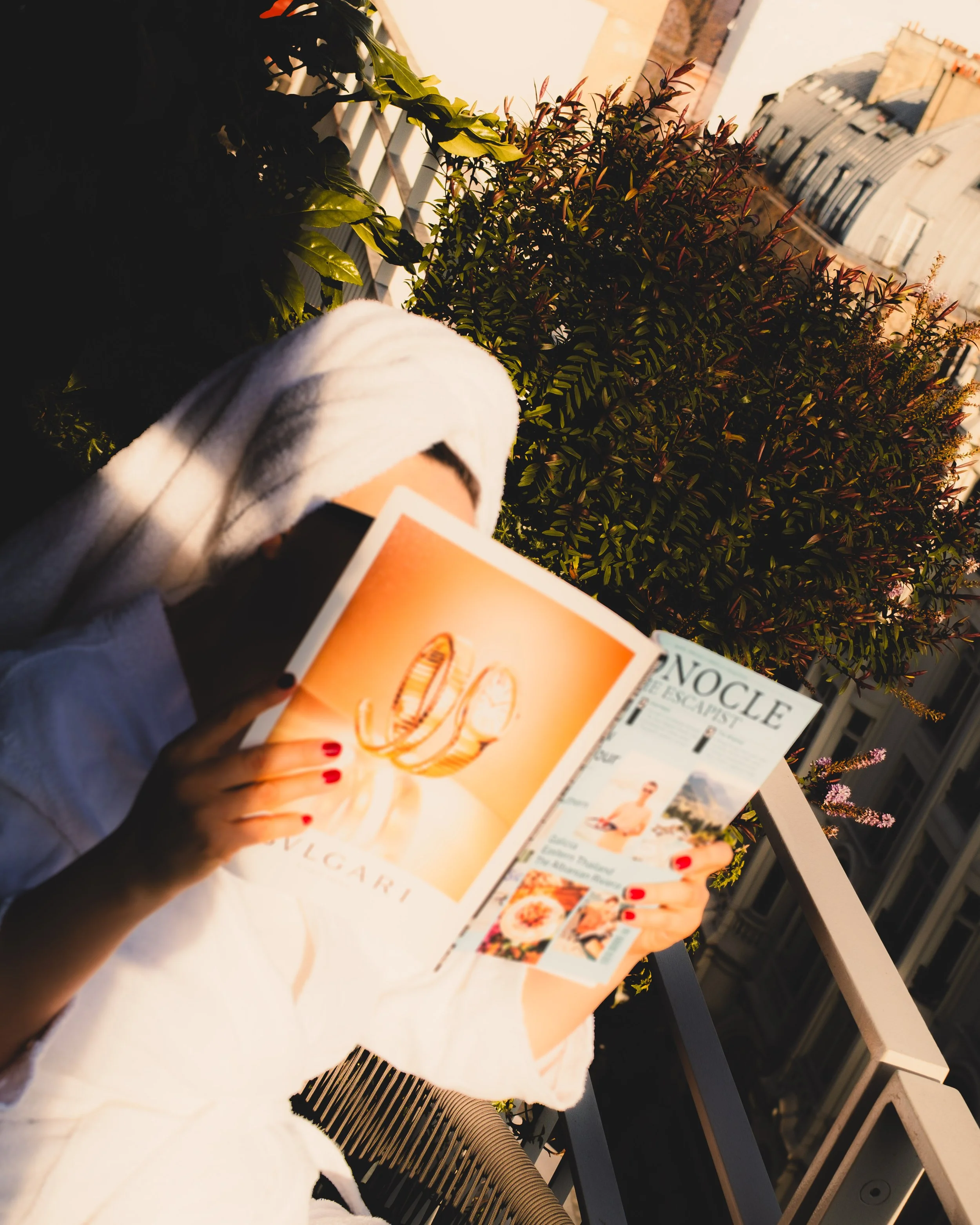 A person holding a magazine with a loaf of bread illustration on the cover. The person has red nail polish and is sitting outdoors at a table with a white tablecloth. There are potted plants and a building with an arched roof in the background.