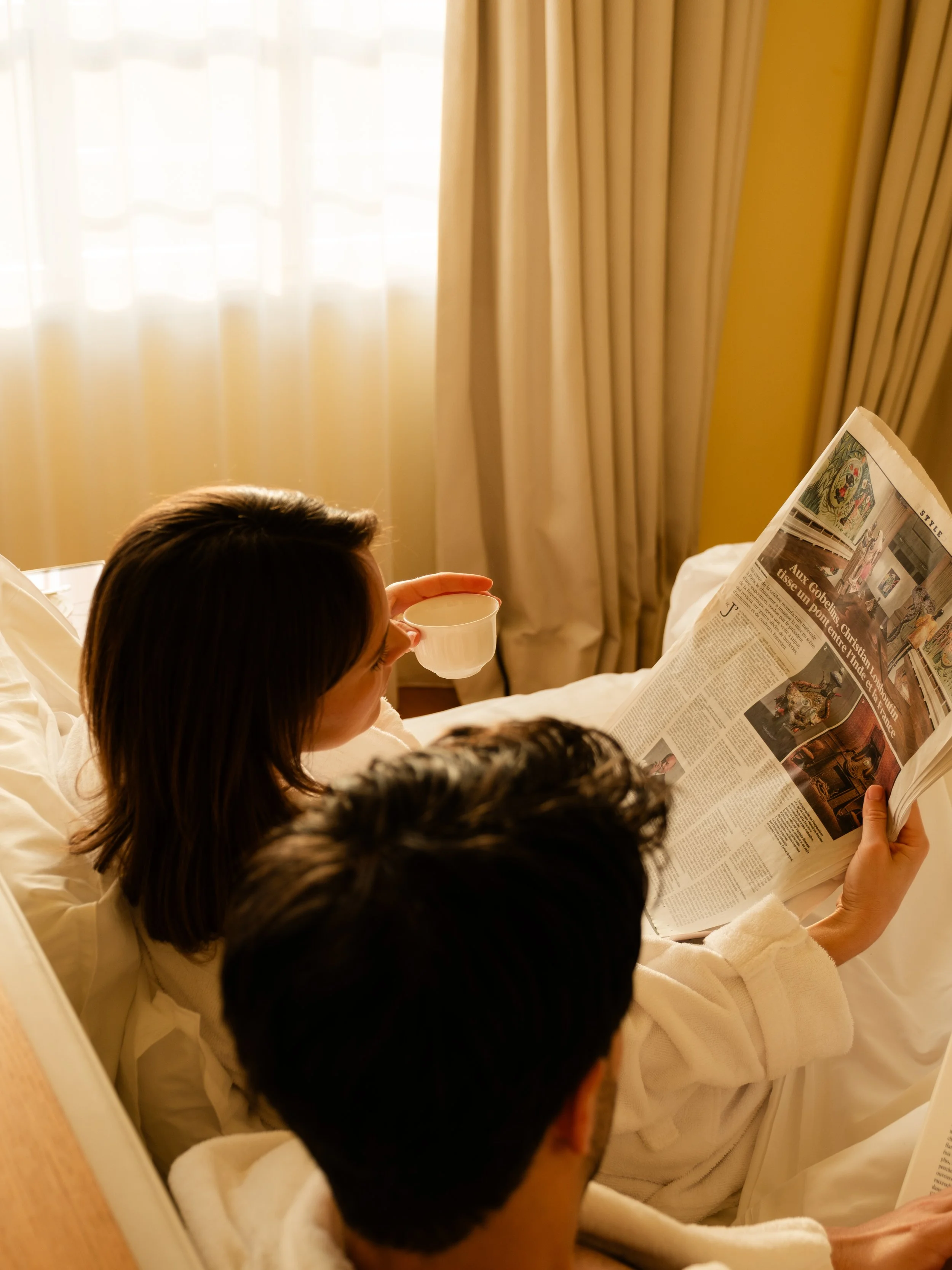 Two people are sitting in bed with white robes, one reading a newspaper and the other holding a cup, in a cozy room with yellow walls and curtains. Lionel Chu - Hotel & Travel Photography