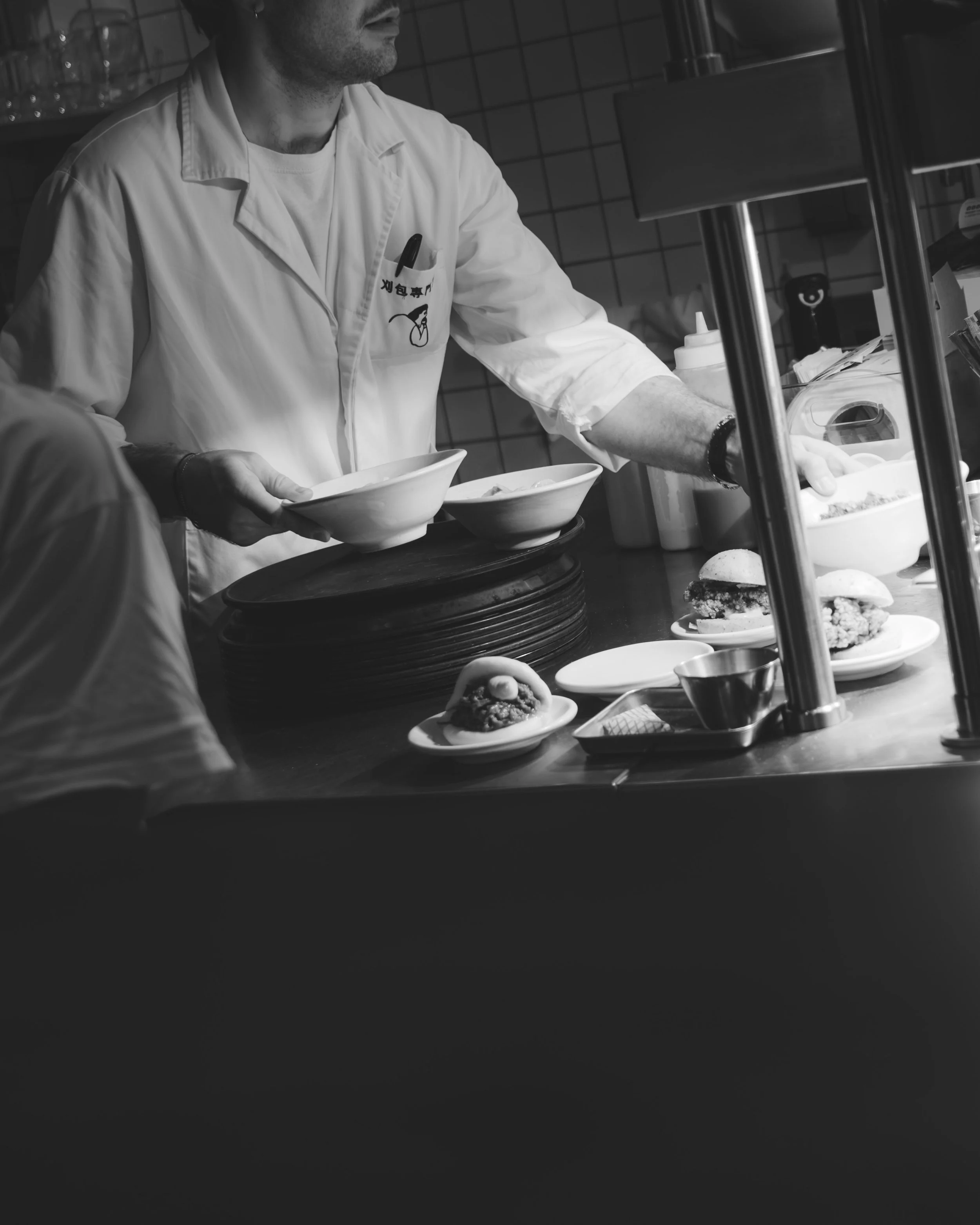 A chef serving food in a restaurant kitchen, holding a bowl while standing next to a food station with plates of food.