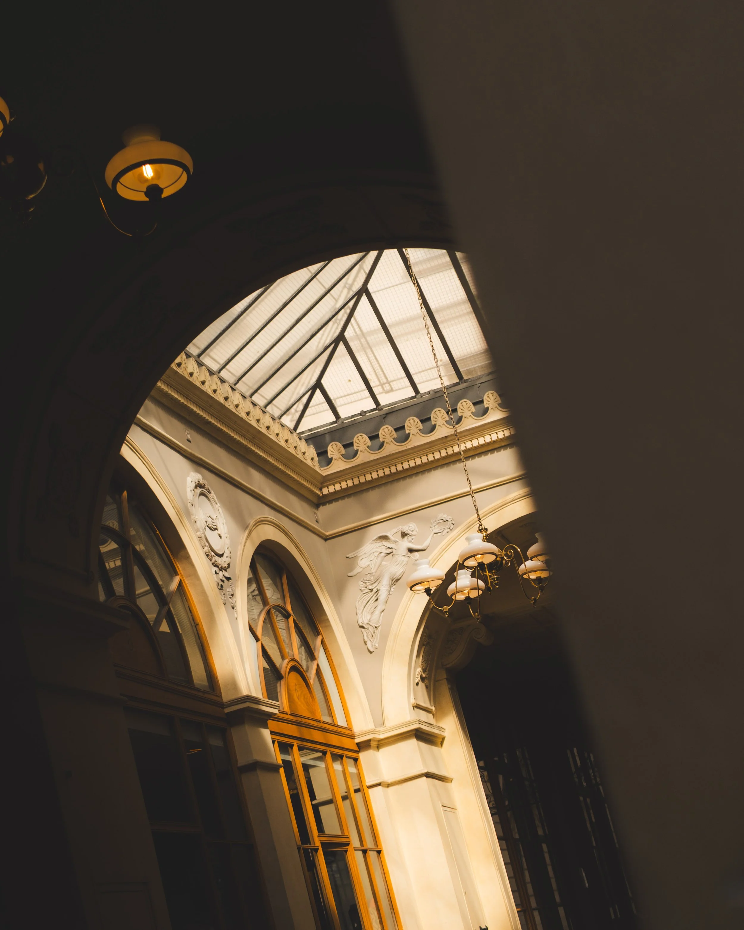 Interior of an ornate building with a curved ceiling, decorative molding, a statue of an angel, and a glass roof allowing natural light to illuminate the space.