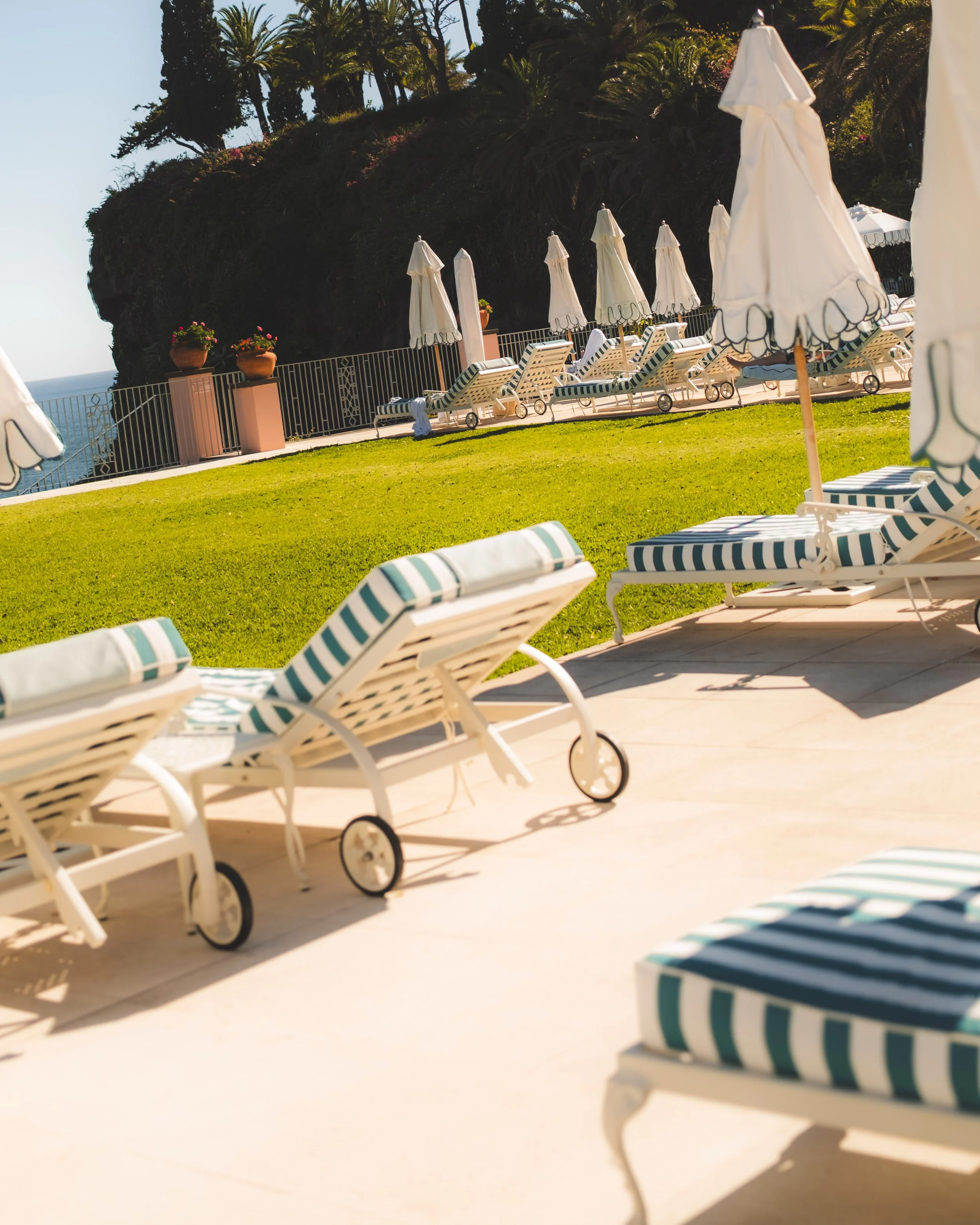 Empty poolside lounge chairs with striped cushions and closed umbrellas on a sunny day, with greenery and trees in the background.