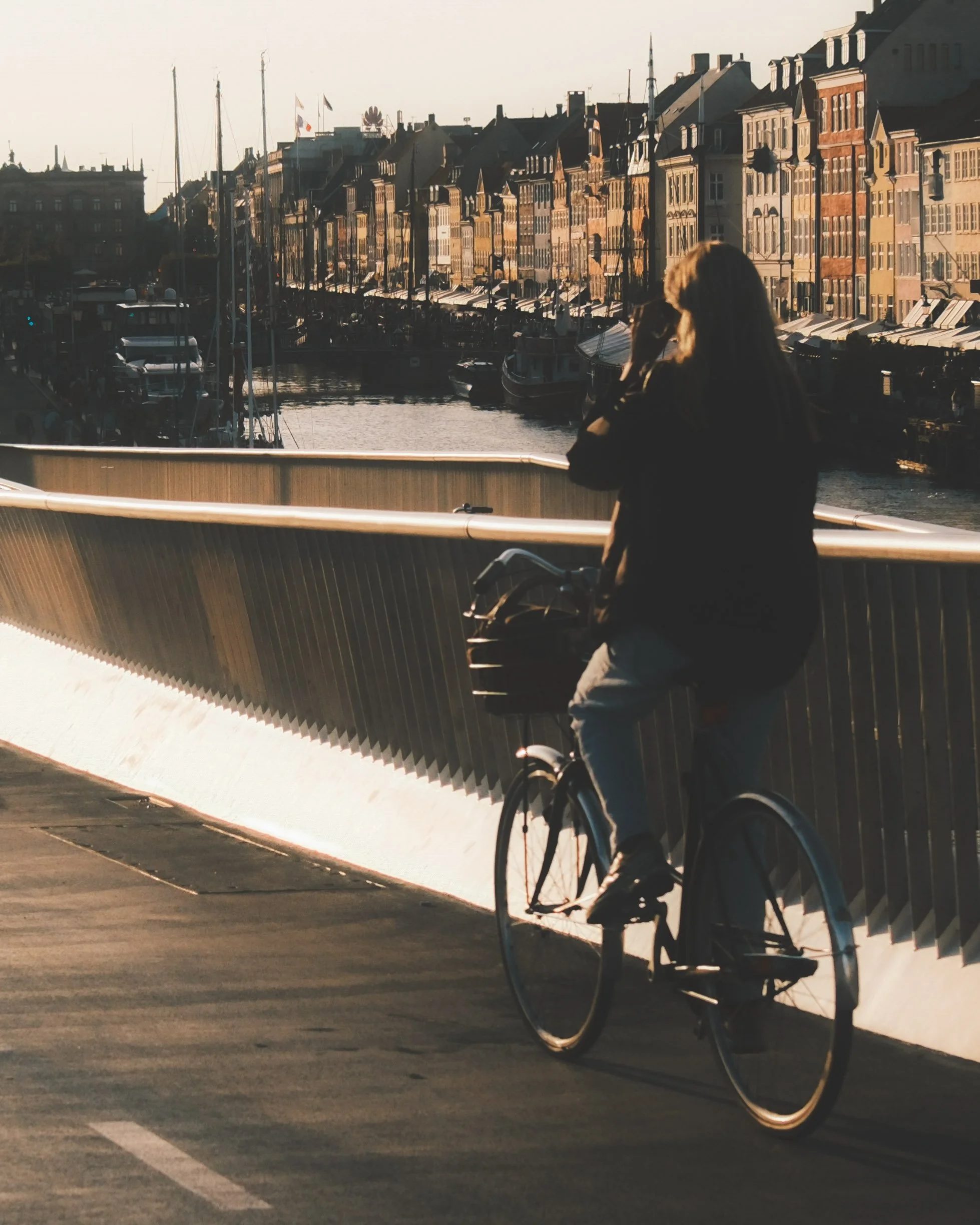 A person with long hair riding a bicycle along a waterfront promenade at sunset, with historic buildings and a marina with boats in the background.