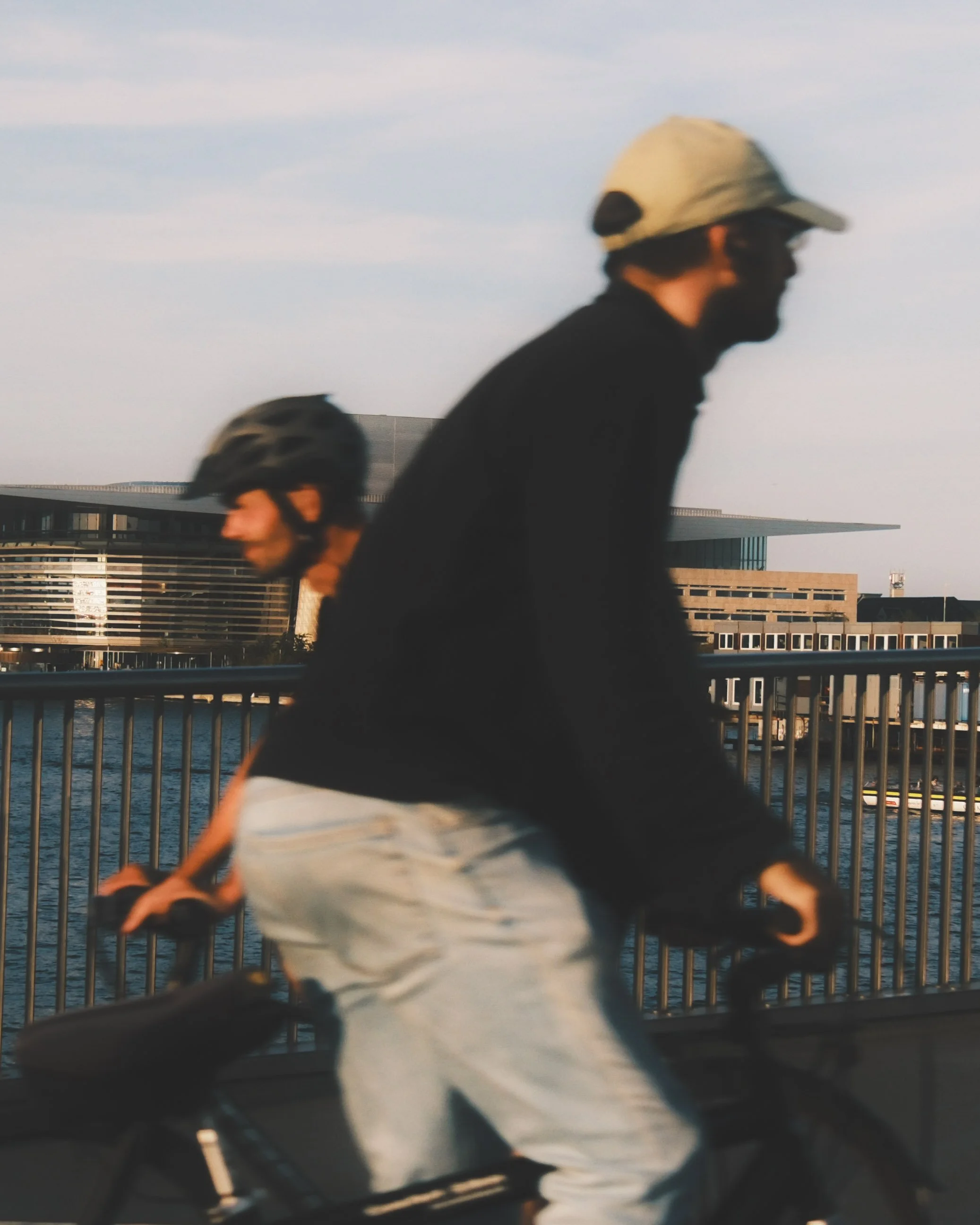 Two people cycling along a bridge during sunset, with modern buildings and a cloudy sky in the background.