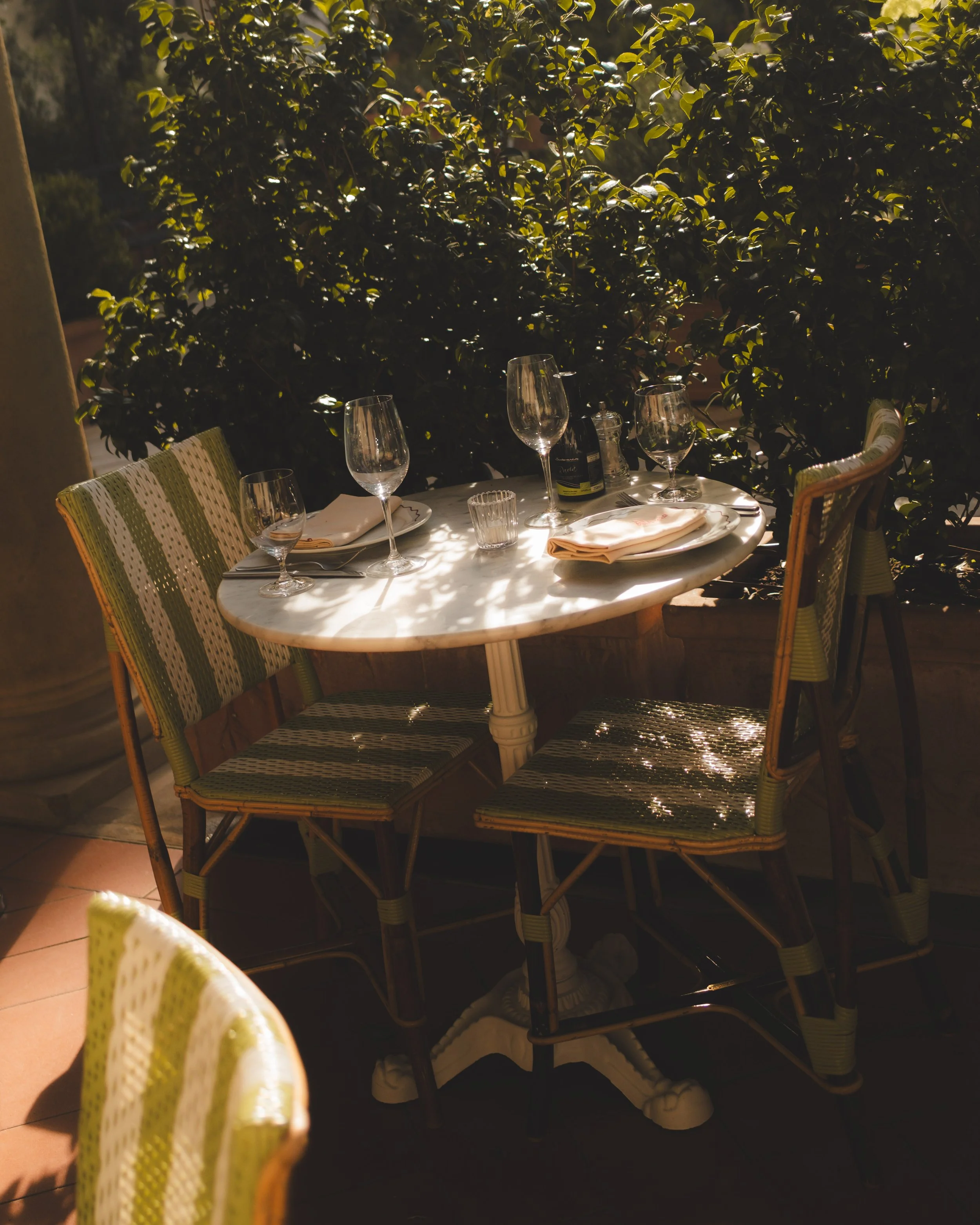 An outdoor dining table set with wine glasses, a small bottle, napkins, and plastic cups, surrounded by wicker chairs with green cushions, under warm sunlight with lush greenery in the background.