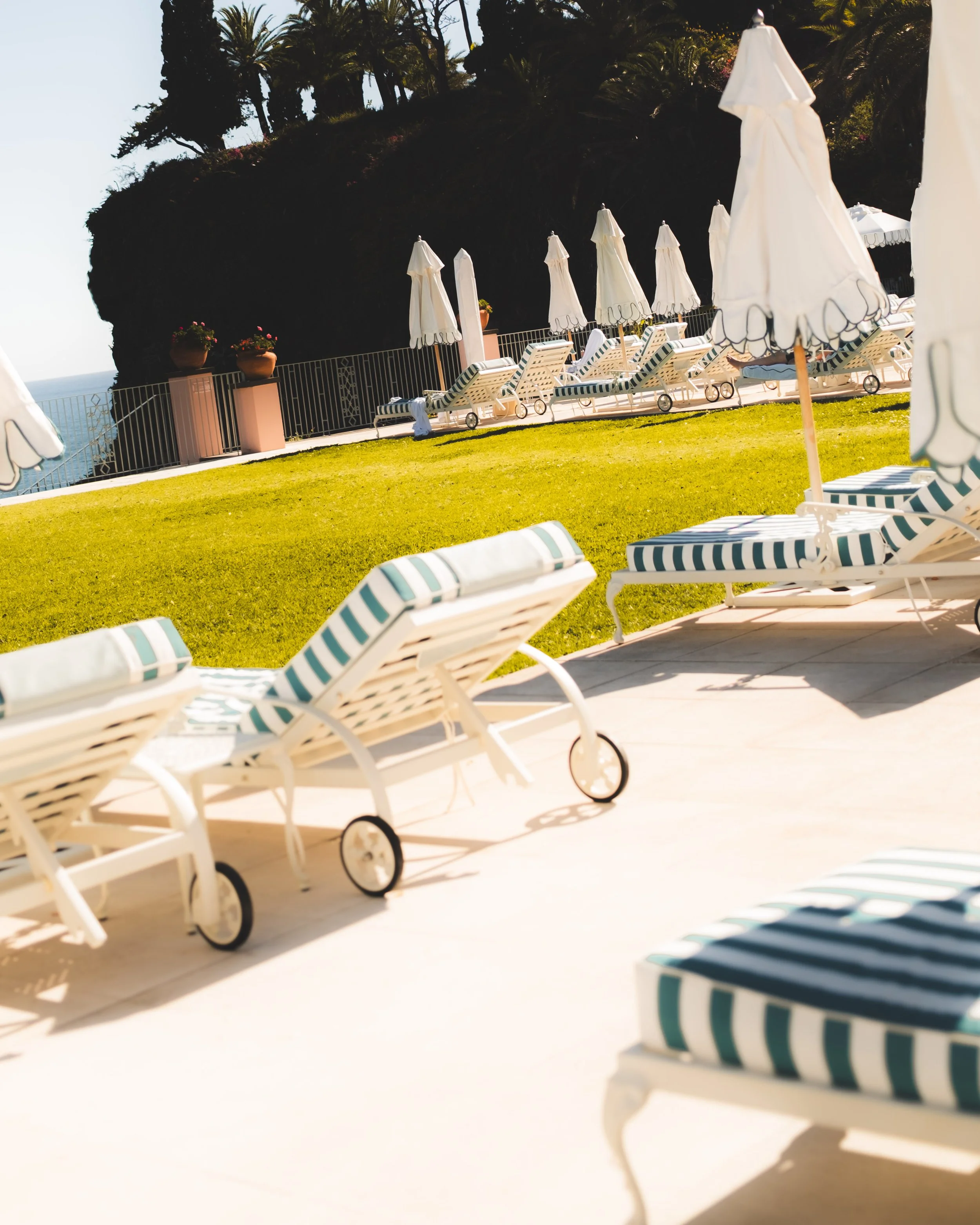 Empty poolside lounge chairs with striped cushions and closed umbrellas on a sunny day, with greenery and trees in the background.