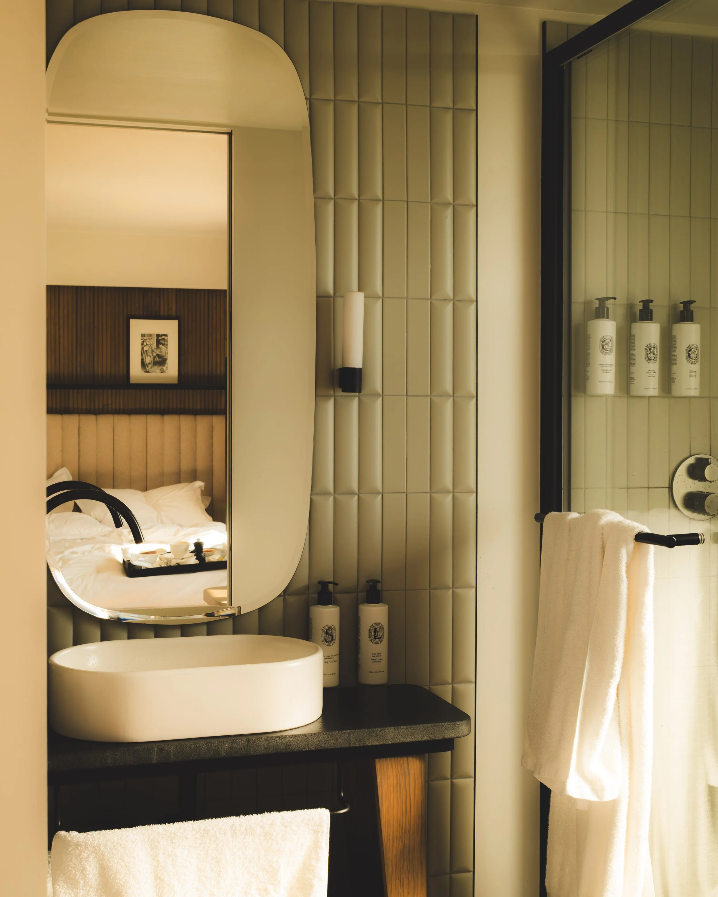 A bathroom vanity area with a round sink, a large mirror, and toiletries including shampoo and lotion bottles placed on a shelf. There is a beige padded wall behind and a white towel hanging on a black towel rack.