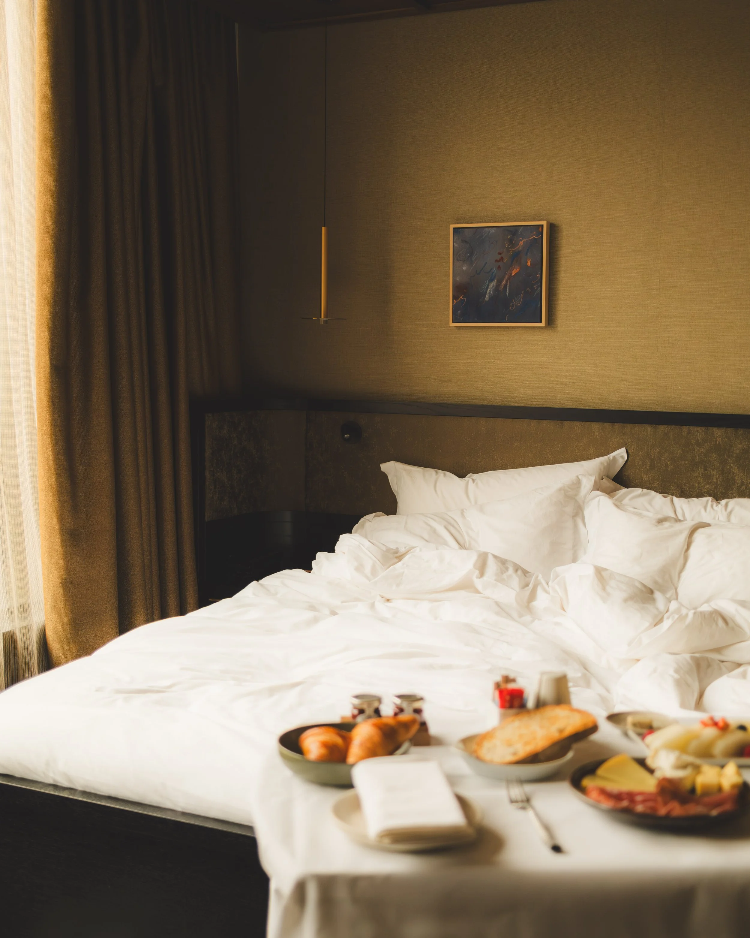 A breakfast tray with food on a bed in a hotel room. The tray includes pastries, eggs, fruits, and condiments. The bed has white linens, and the headboard behind has a minimalist wall decor and a hanging light fixture. There are curtains on the left side of the room.