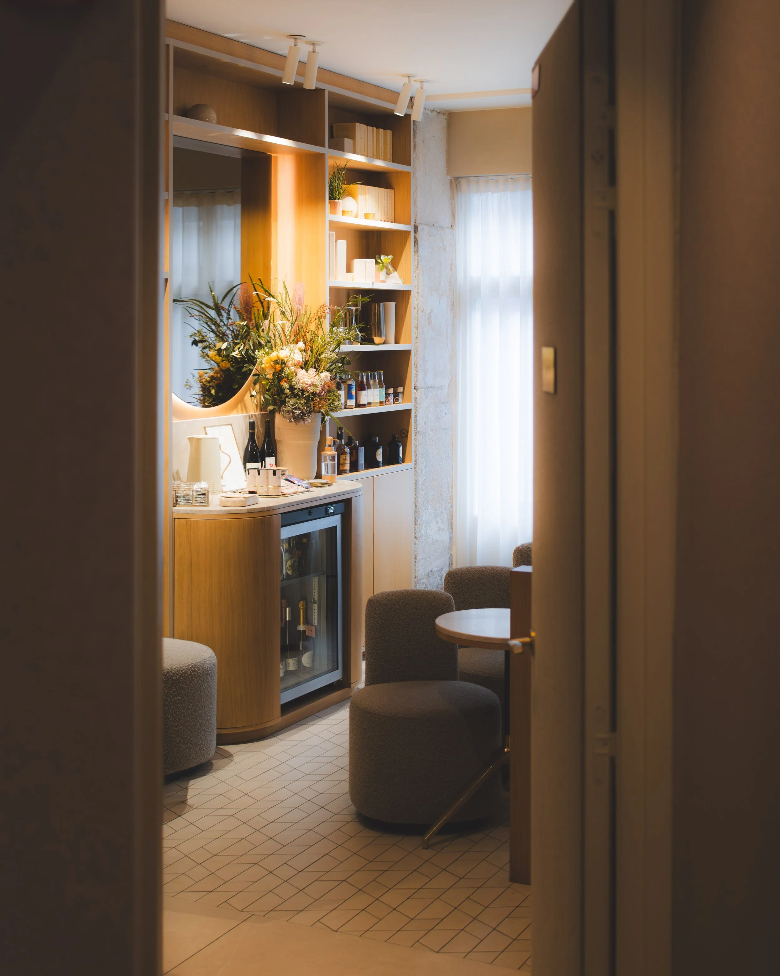 Interior of a cozy café or bar with a wooden shelving unit holding books, plants, and decorative items. There are tables and cushioned chairs with a tiled floor, and sunlight filters through sheer curtains on the windows.