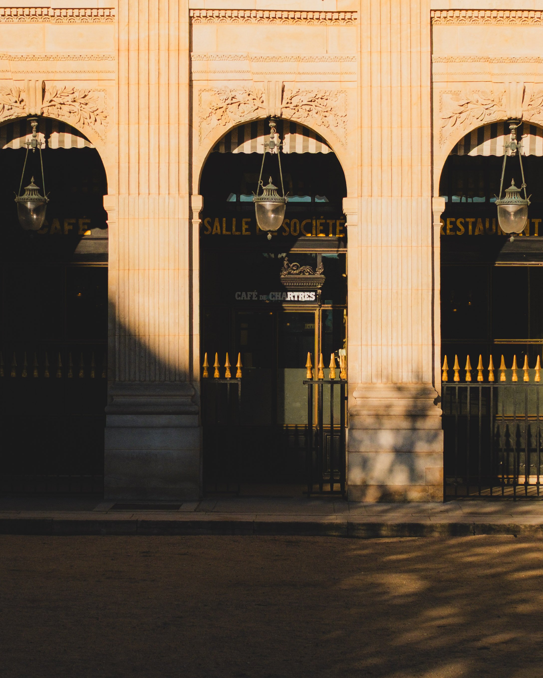 Facade of a historic building with arched doorways, decorative columns, ornate carvings, and hanging lanterns. A sign reads 'Café de Chartres.' Palais Royal garden. Lionel Chu - Hotel & Travel Photographer.