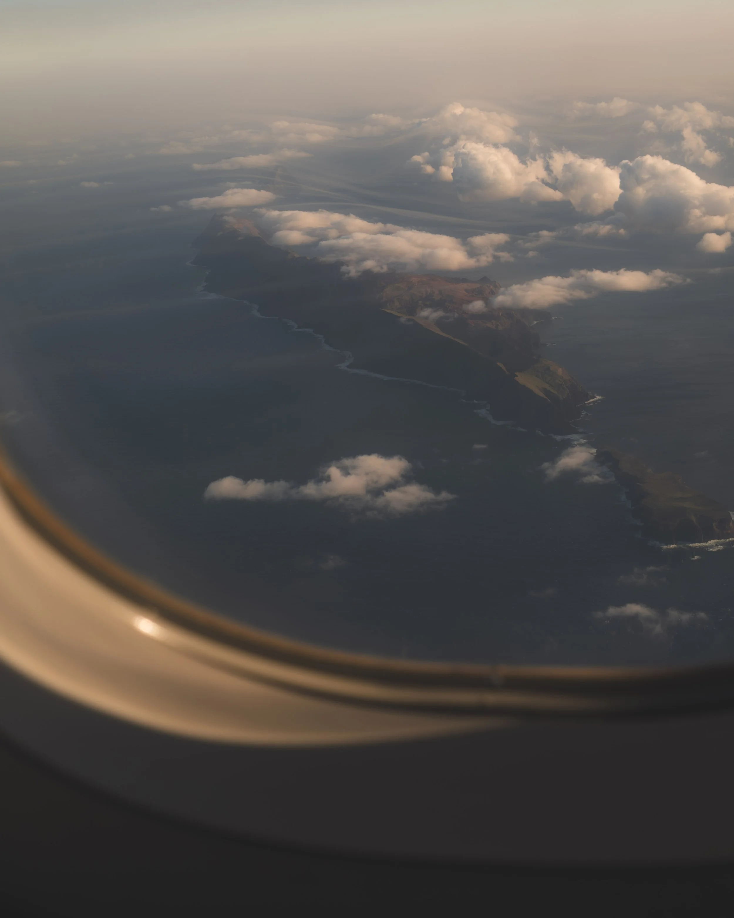 An aerial view of a coastline with rugged cliffs, green patches, and stormy clouds over the sea, seen from an airplane window.