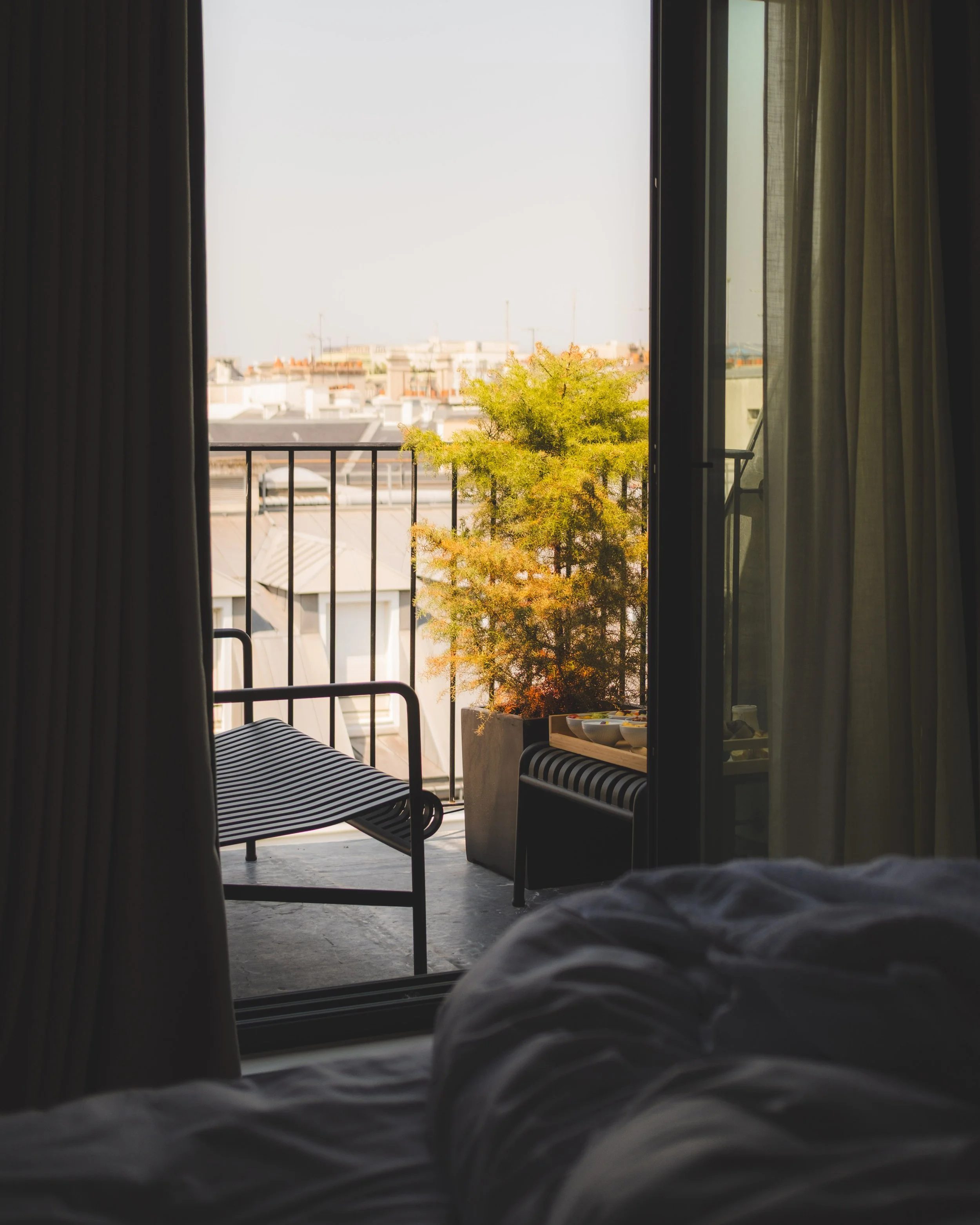 Photo taken from a bed showing a balcony with a black metal railing, a yellow-green tree in a planter, and a cityscape in the background through an open glass door, with curtains on either side.