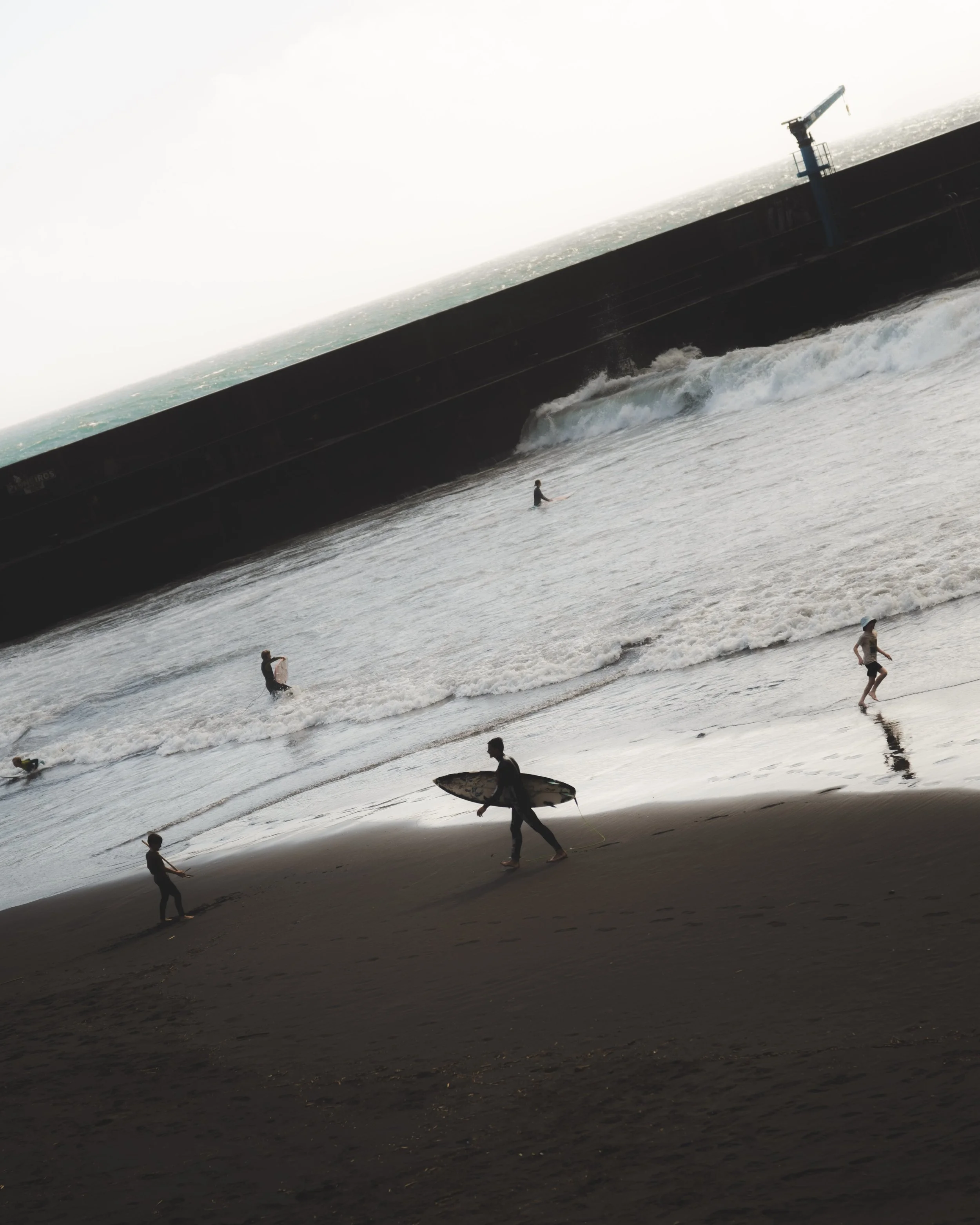 People at the beach, some surfing, some walking, with waves and a large ship or barge in the ocean in the background, and a hazy sky.
