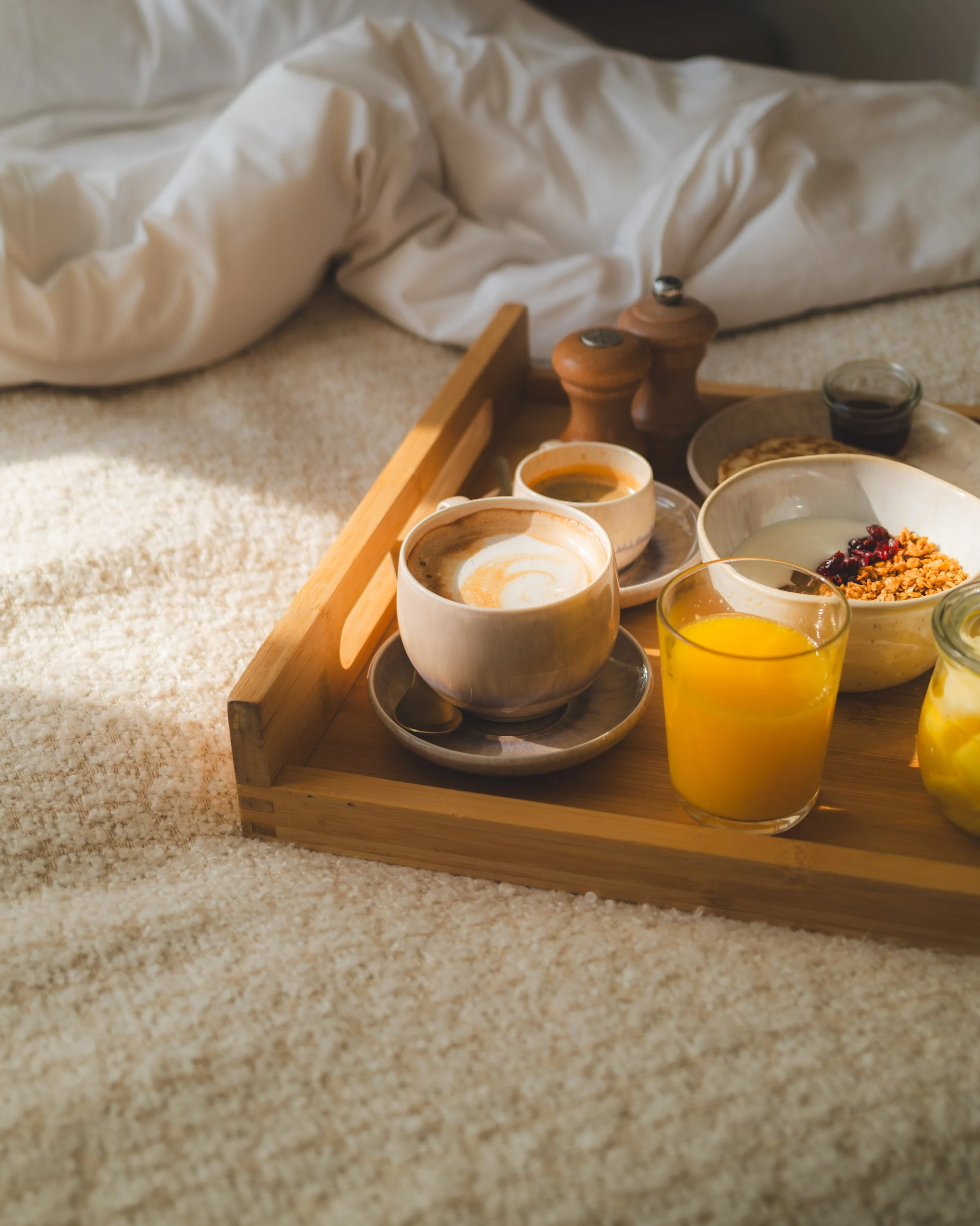 A breakfast tray with coffee, juice, and various bowls on beige carpeting, with a rumpled white bedsheet in the background.