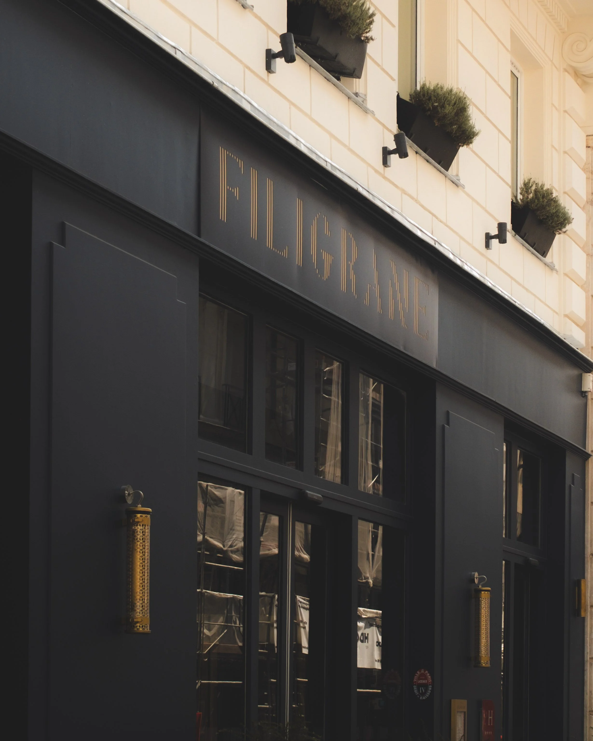 The image shows the exterior of a building with a black sign that has the word 'FILIGRANE' in gold lettering. Above the sign, there are three flower boxes with greenery on the windowsill, attached to the pale yellow facade.