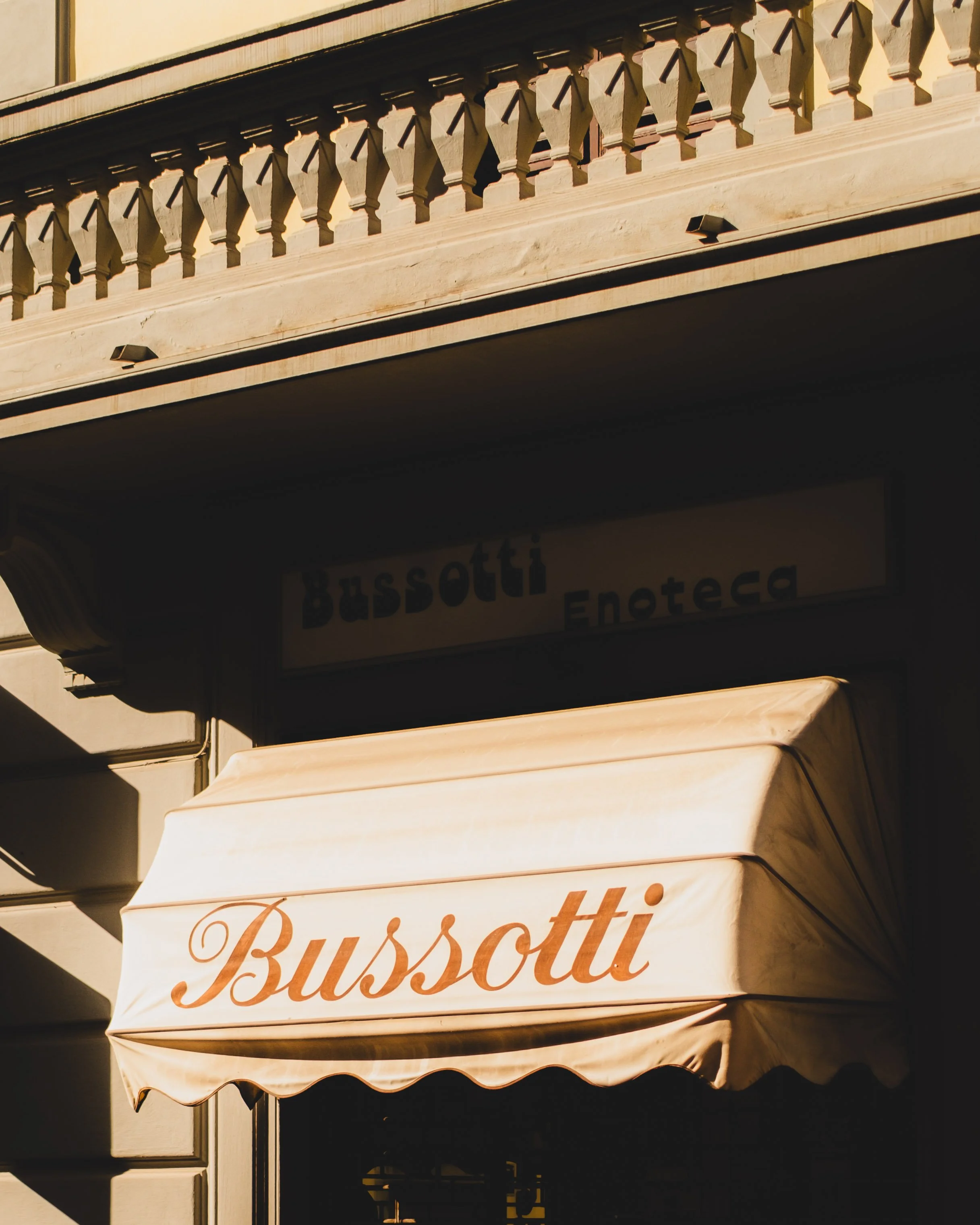 Storefront of Bussotti Enoteca with a beige awning displaying the name 'Bussotti' in brown cursive lettering, set outside a building with a decorative balcony above. Lionel Chu - Hotel & Travel Photographer