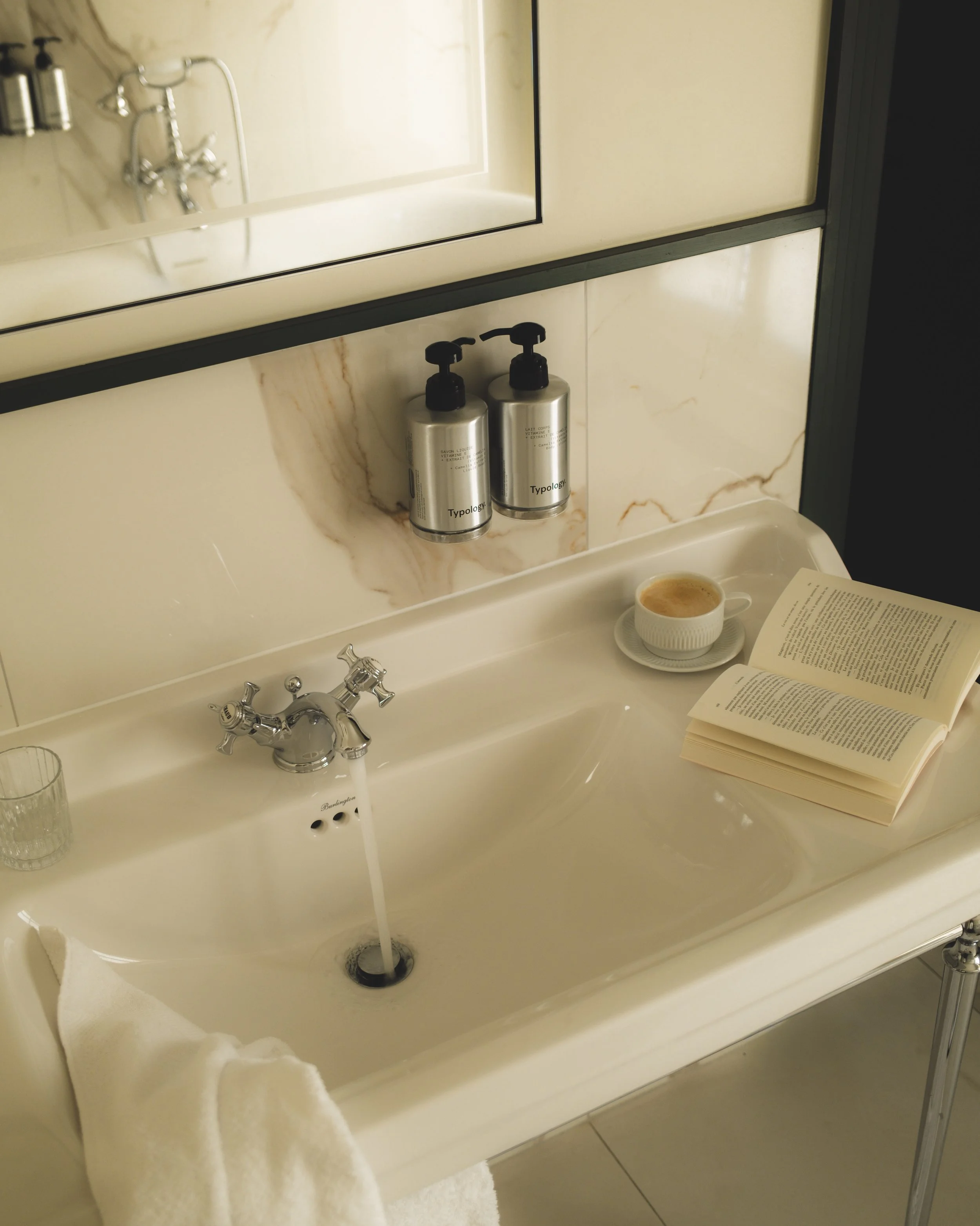 Bathroom sink with a mirror above and two soap dispensers labeled 'Typology.' A cup with a beverage, an open book, and a towel are on the sink.