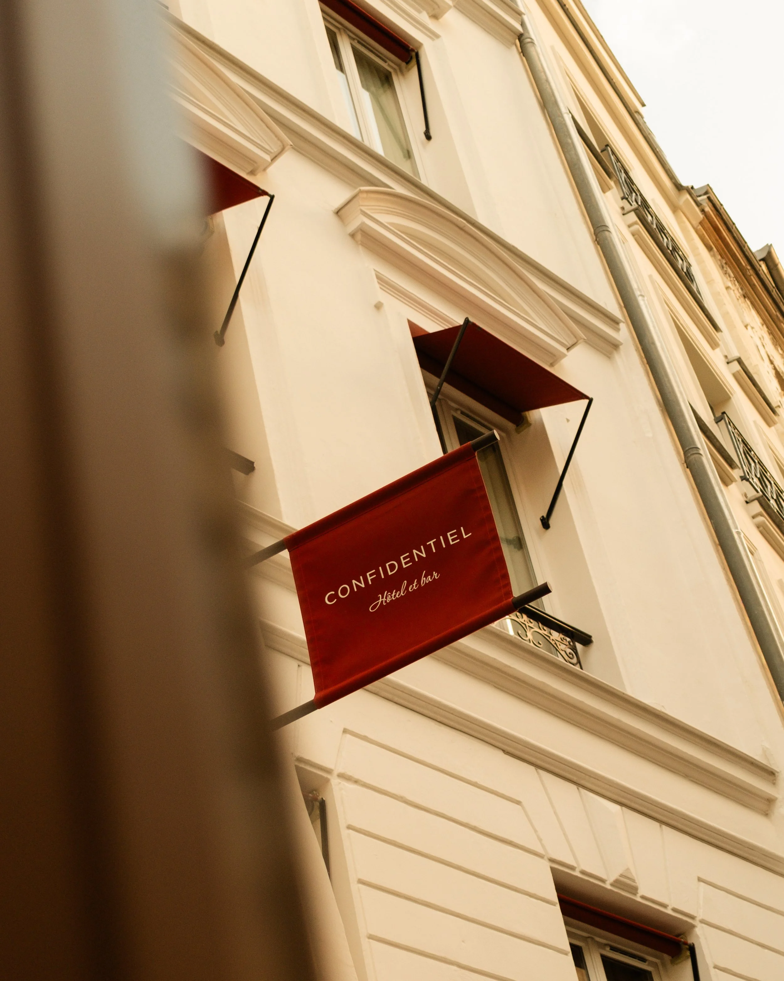 A hotel sign reading "CONFIDENTIEL Hotel et bar" hanging outside a white building with windows and red awnings. Lionel Chu - Hotel & Travel Photography