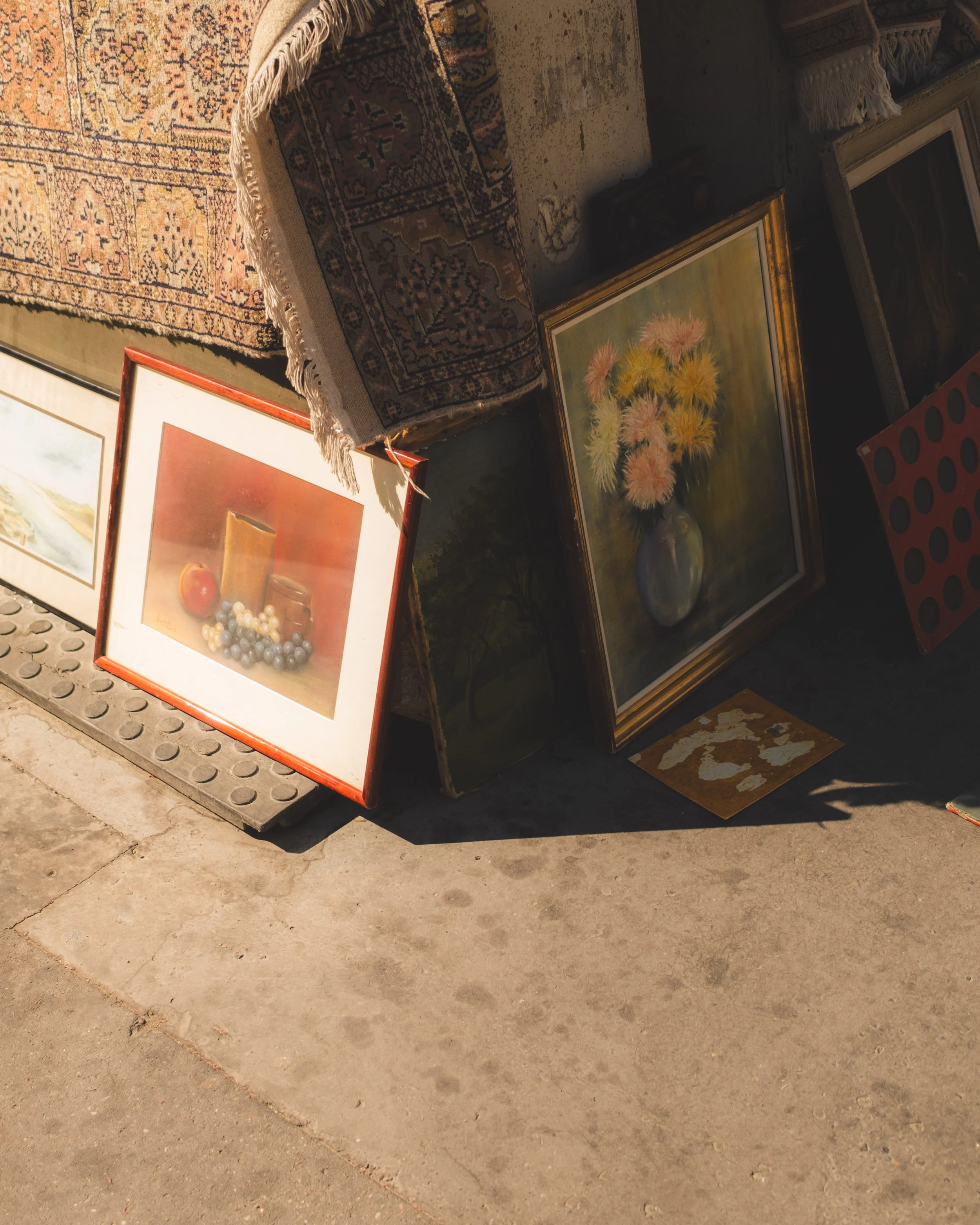 Various framed paintings and rugs leaning against a wall on a concrete sidewalk.