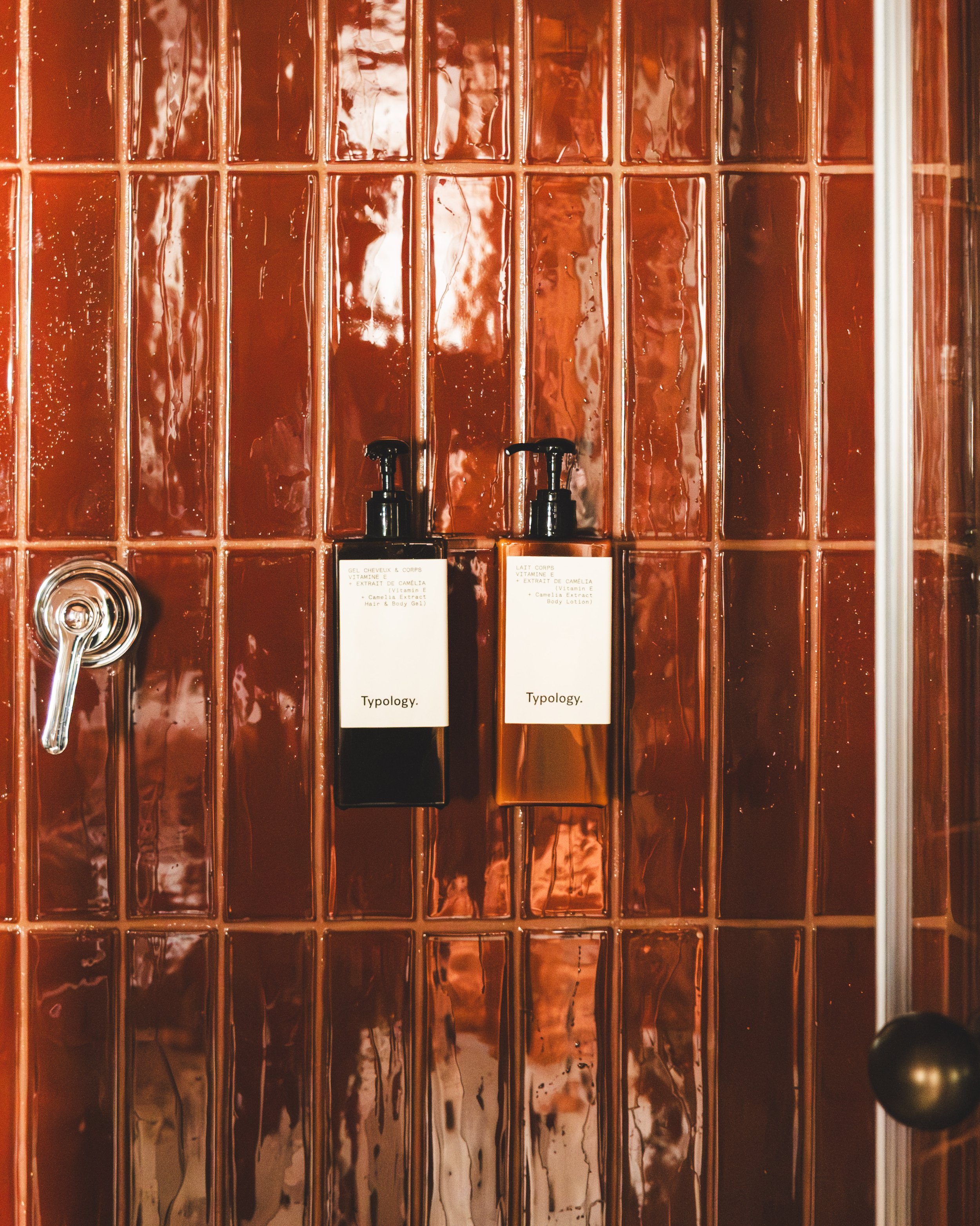 Close-up of copper colored glass tiles in a grid pattern on a wall with a dark soap dispenser and product bottles labeled 'typlogy' in front.