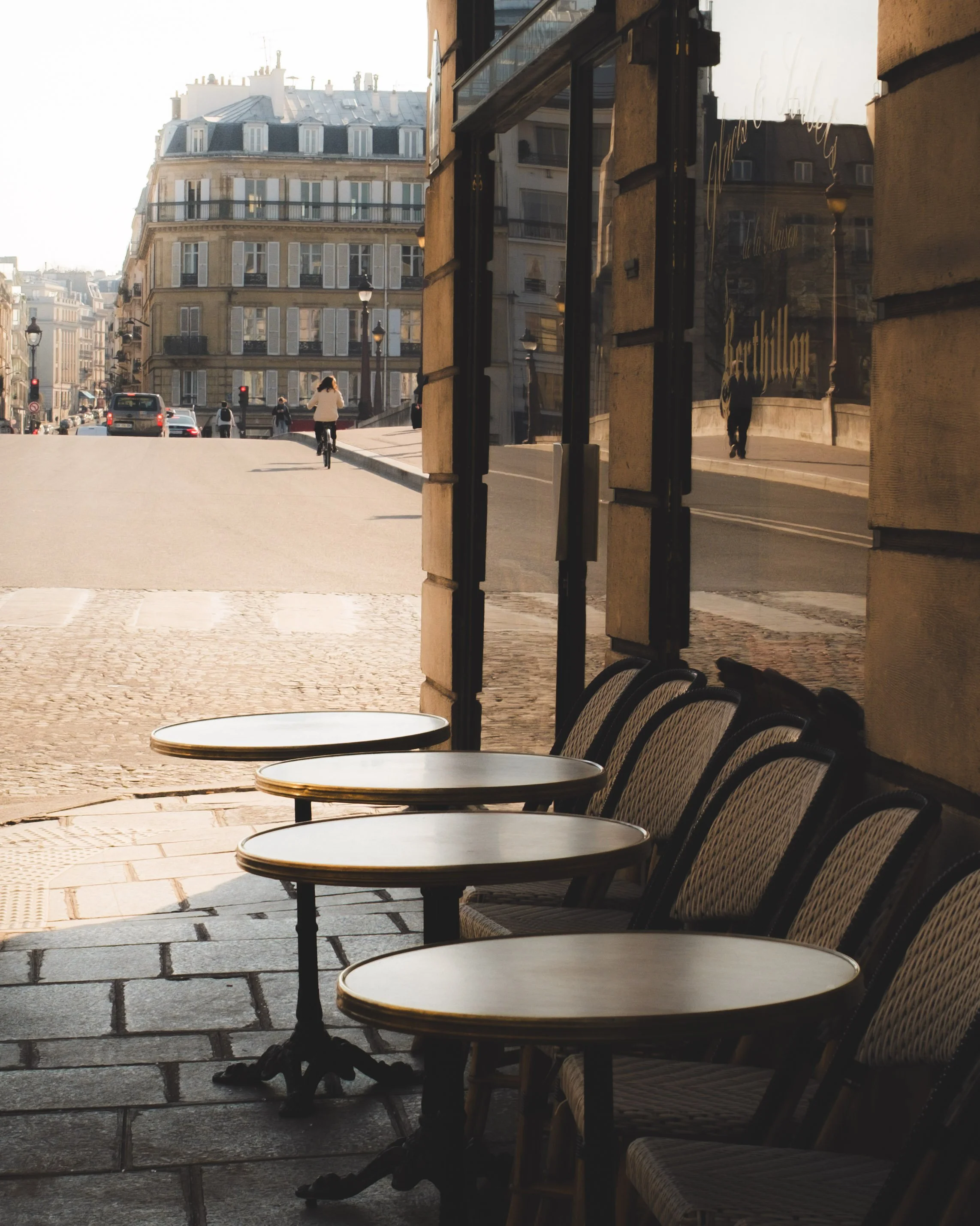 Empty outdoor café tables and chairs are aligned on a sidewalk near a building. The scene captures a sunny day with a street and a cyclist riding away, and buildings in the background reflecting in a window.