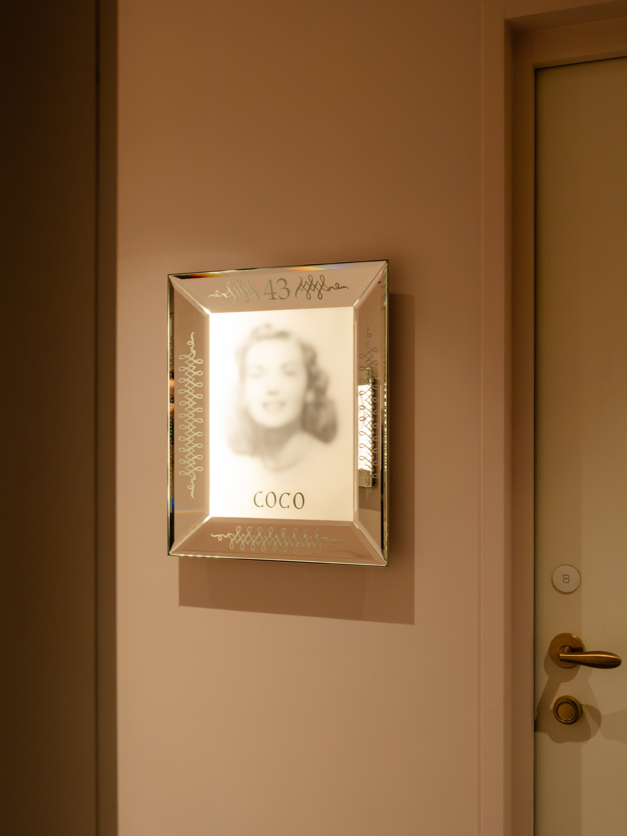 Framed black-and-white portrait of Marilyn Monroe, with the name 'Coco' and the number '43' written on the frame, mounted on a beige wall near a door of an hotel. Lionel Chu - Hotel & Travel Photography