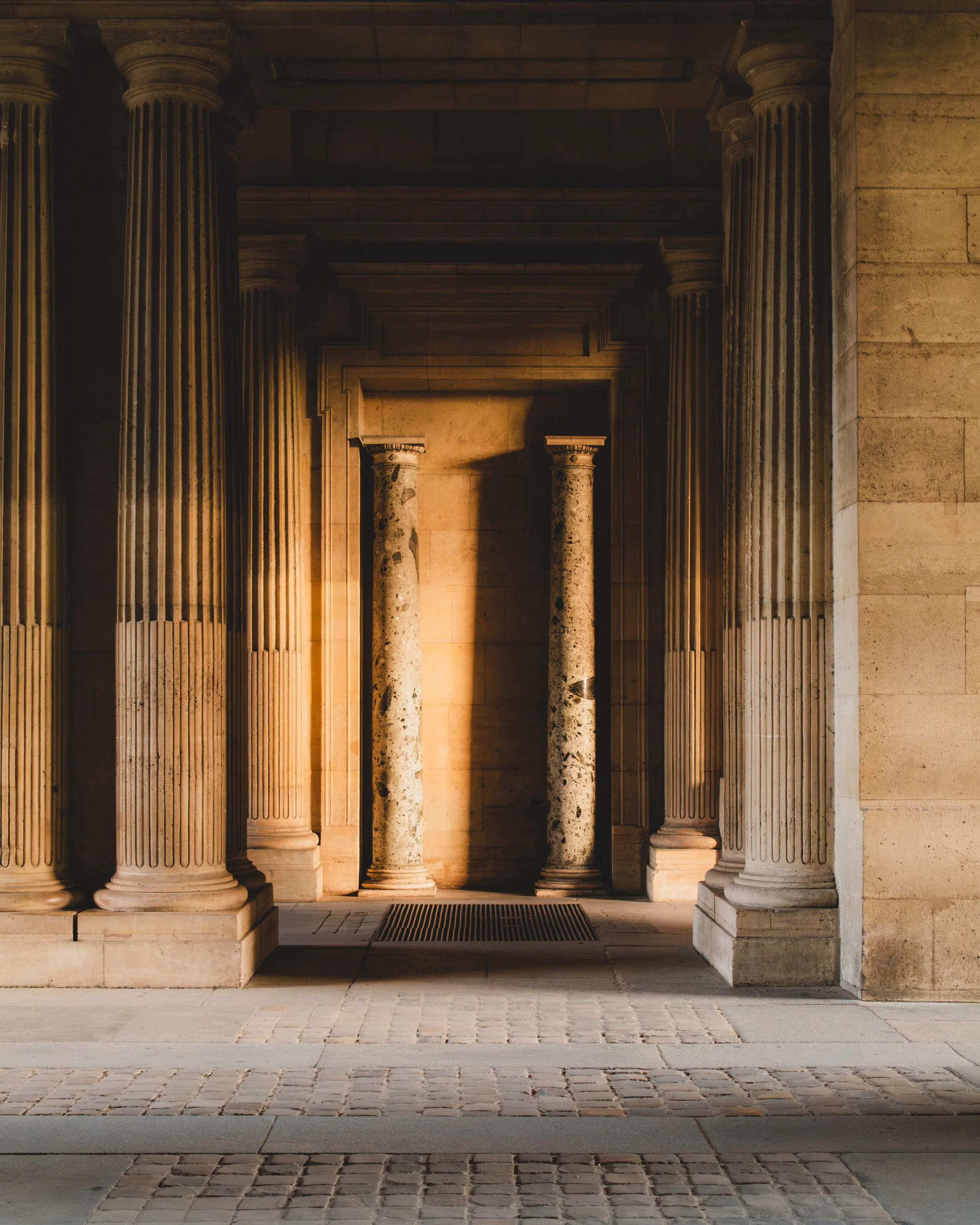 Ancient Greek or Roman-style stone columns with a shadowed corridor behind them, under warm lighting, in Paris, close to the Louvre Museum. Lionel Chu - Hotel & Travel Photographer