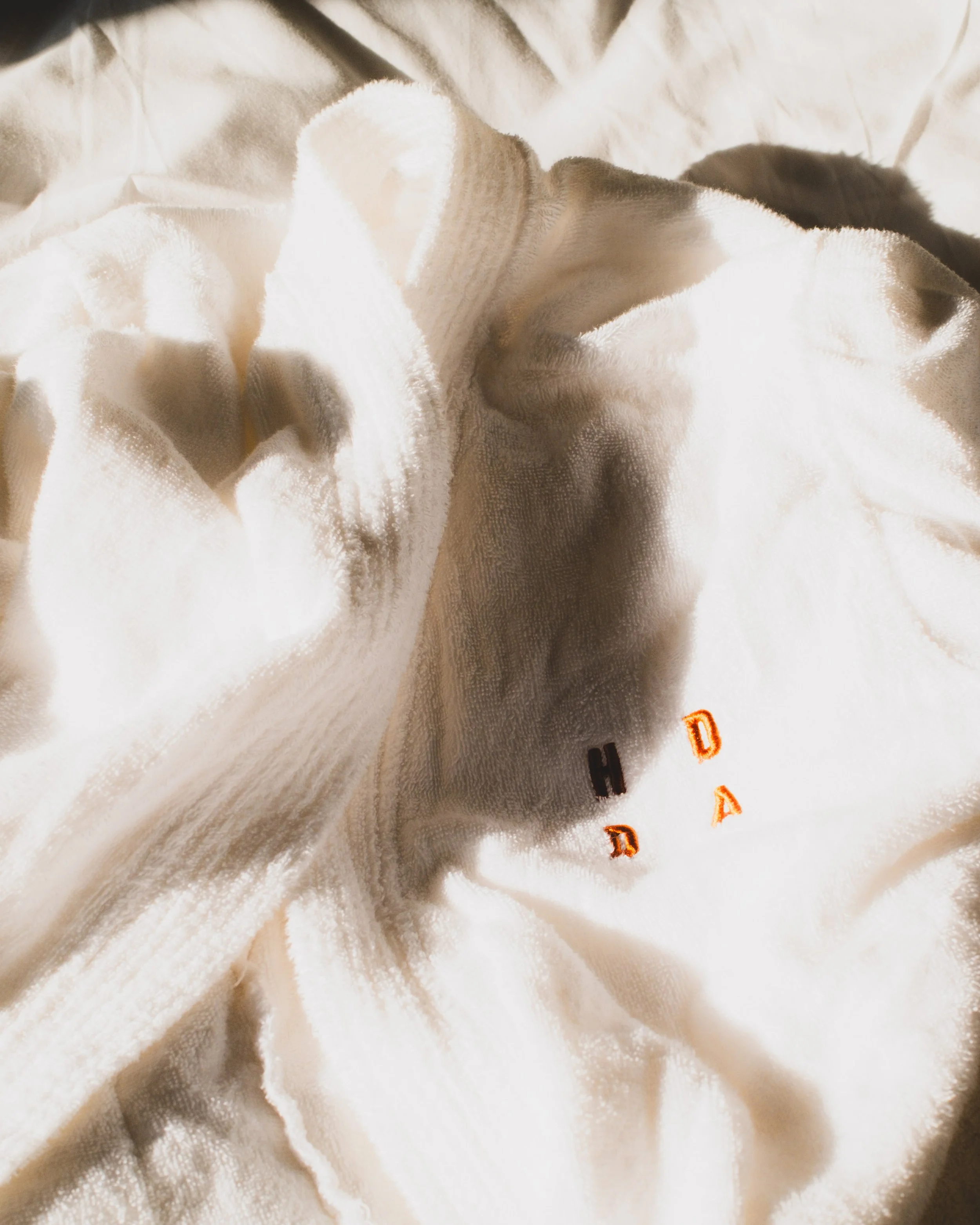 Close-up of soft, cream-colored fabric with broderie anglaise or embroidered detailing. The fabric appears plush and textured, with some visible folds and fraying edges. A small embroidered or printed detail in black and orange is partially visible at the bottom right corner.