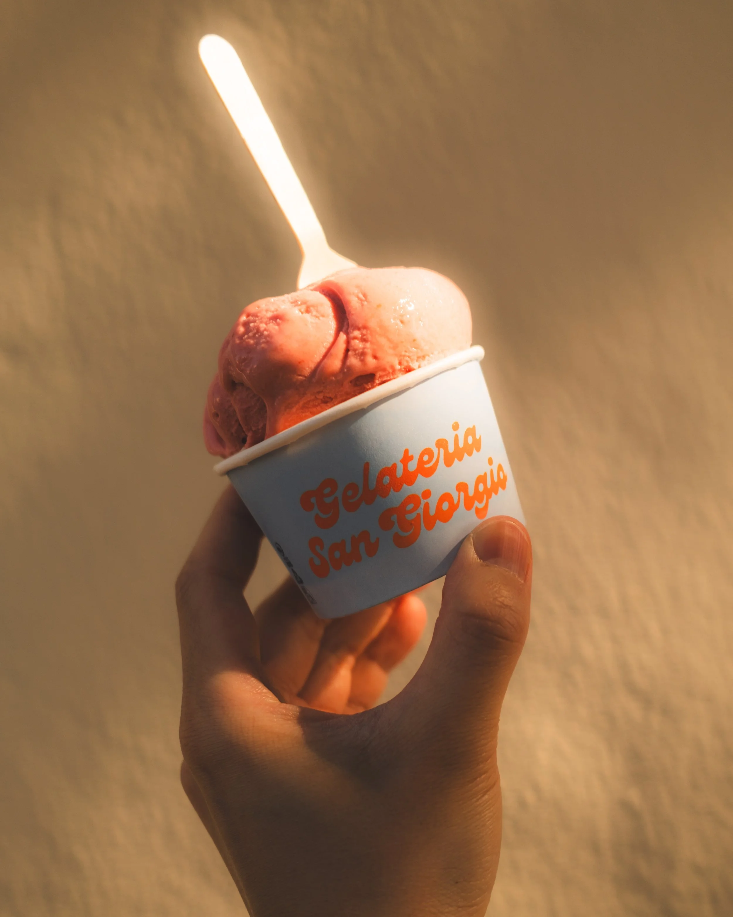 Person holding a cup of strawberry gelato with a lit matchstick inserted into the gelato.