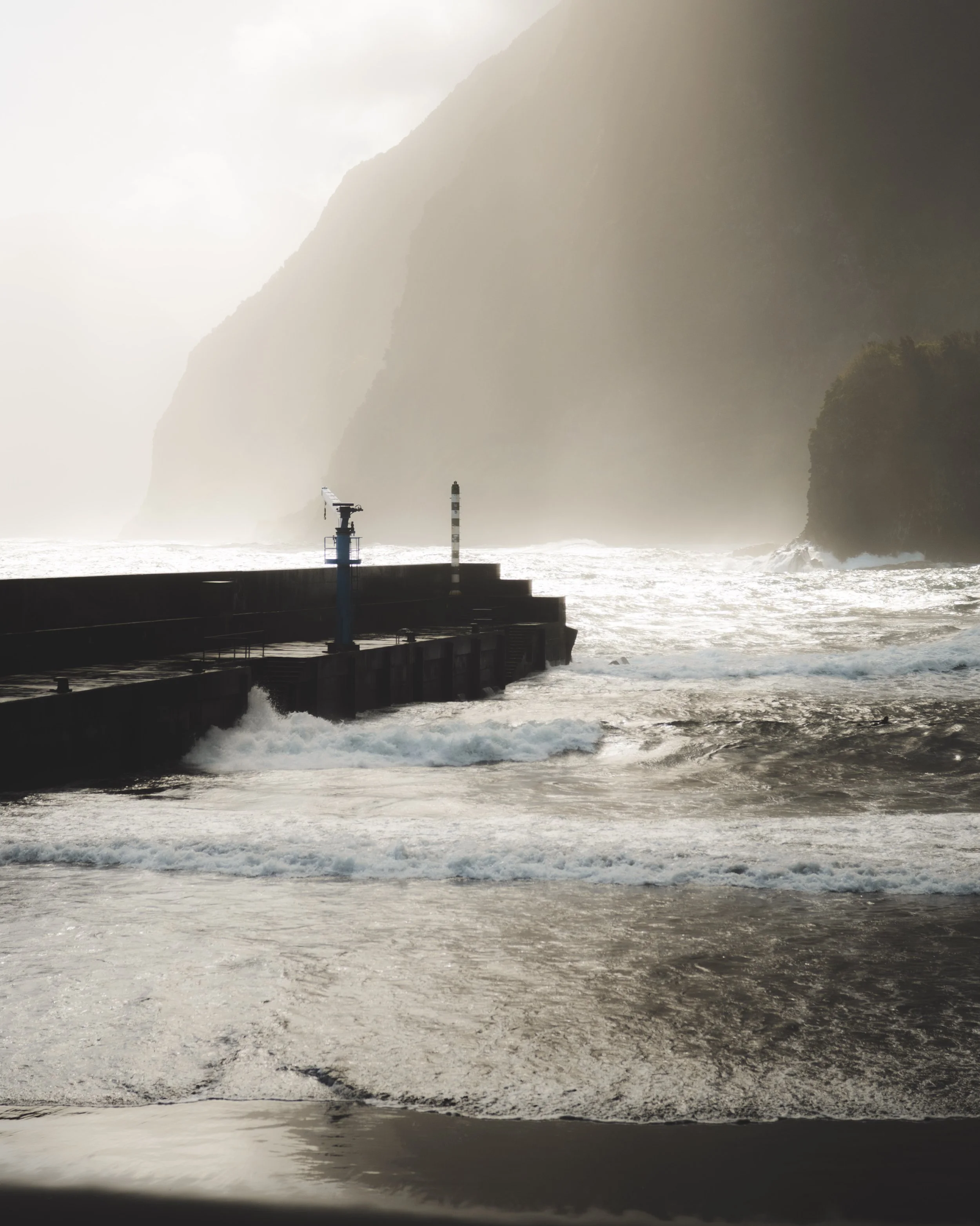 A coastal scene with a breakwater pier, a warning light, large waves, and a foggy atmosphere with mountains in the background.