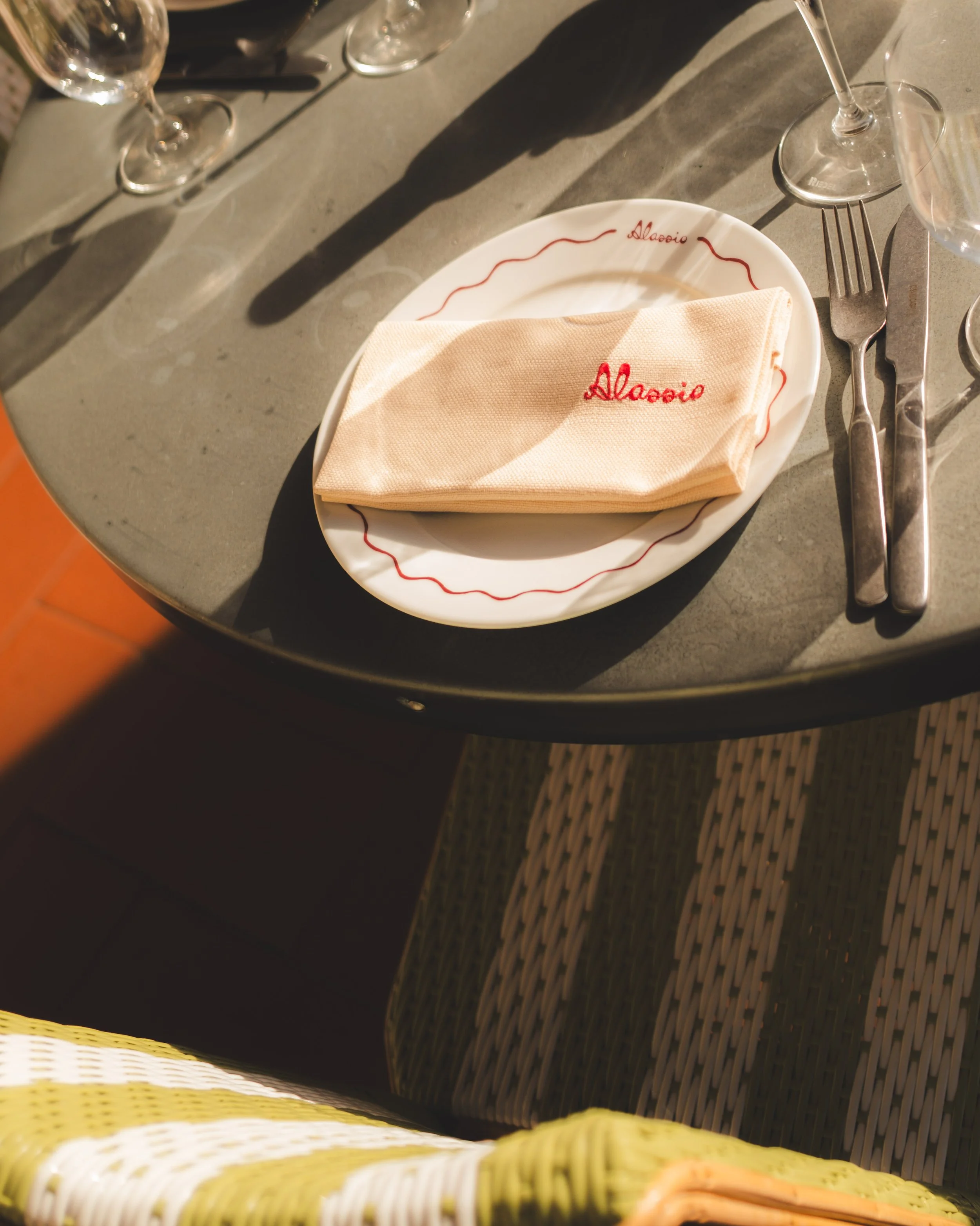 Table setting with wine glasses, a plate with a folded napkin embroidered with 'Alappio,' and cutlery, with sunlight casting shadows on the table.