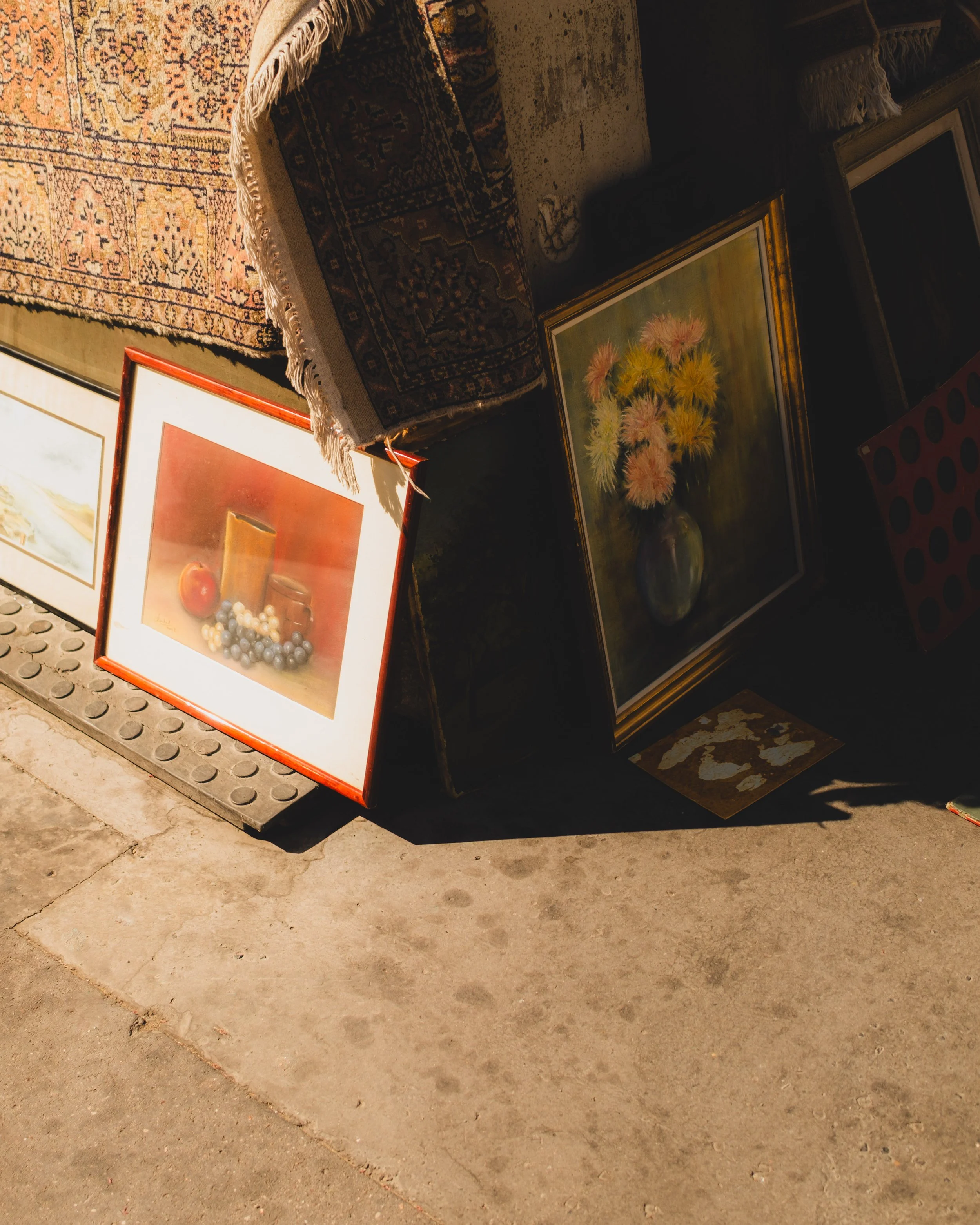 A sidewalk display of framed paintings leaning against a wall, with part of a patterned rug on top. The paintings include still life with candles and fruit, a landscape, and a bouquet of flowers in a vase. The surface is concrete, with sunlight casti