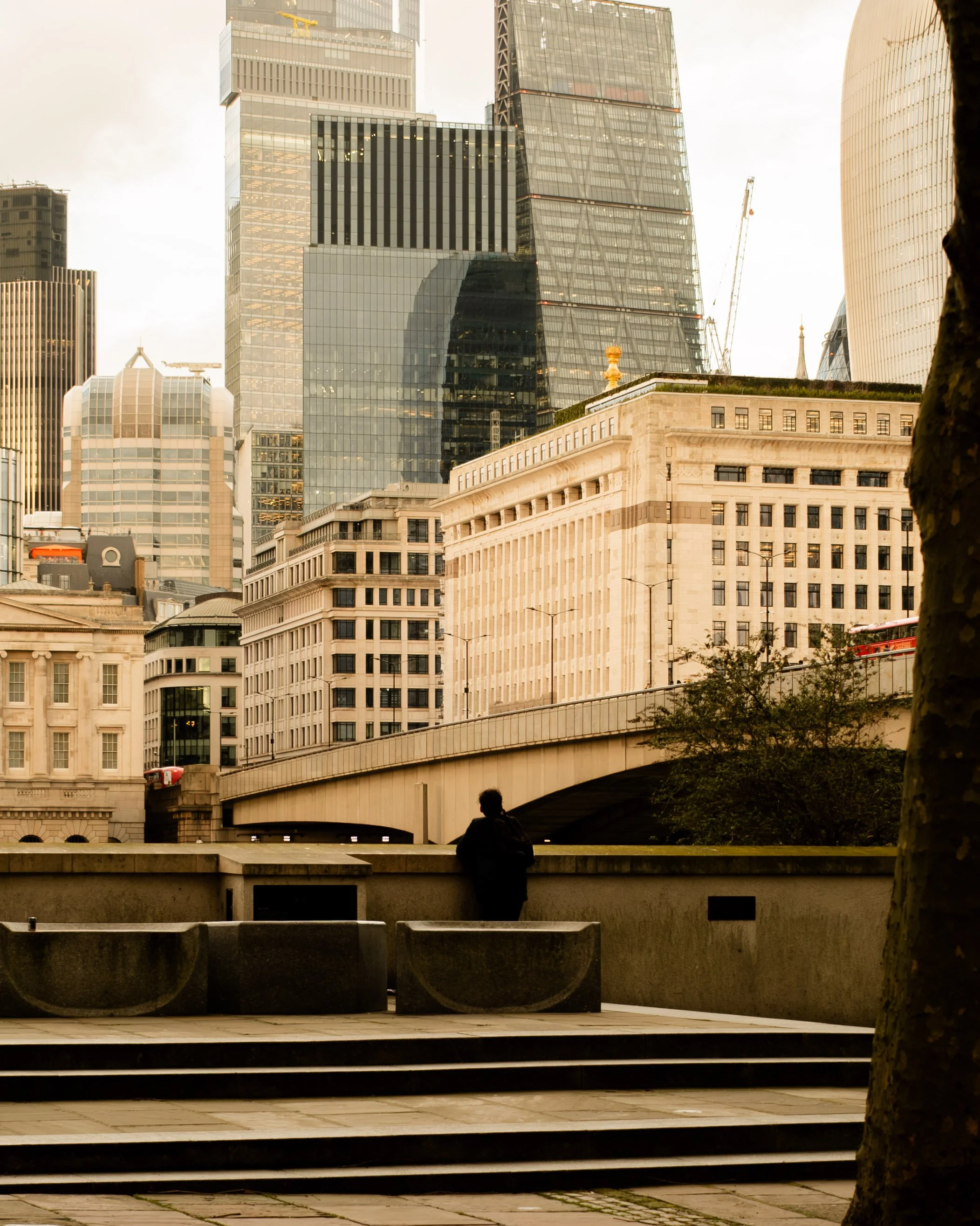 A person standing alone on a city sidewalk with tall modern skyscrapers and historic buildings in the background, during daylight hours. Lionel Chu - Hotel & Travel Photographer