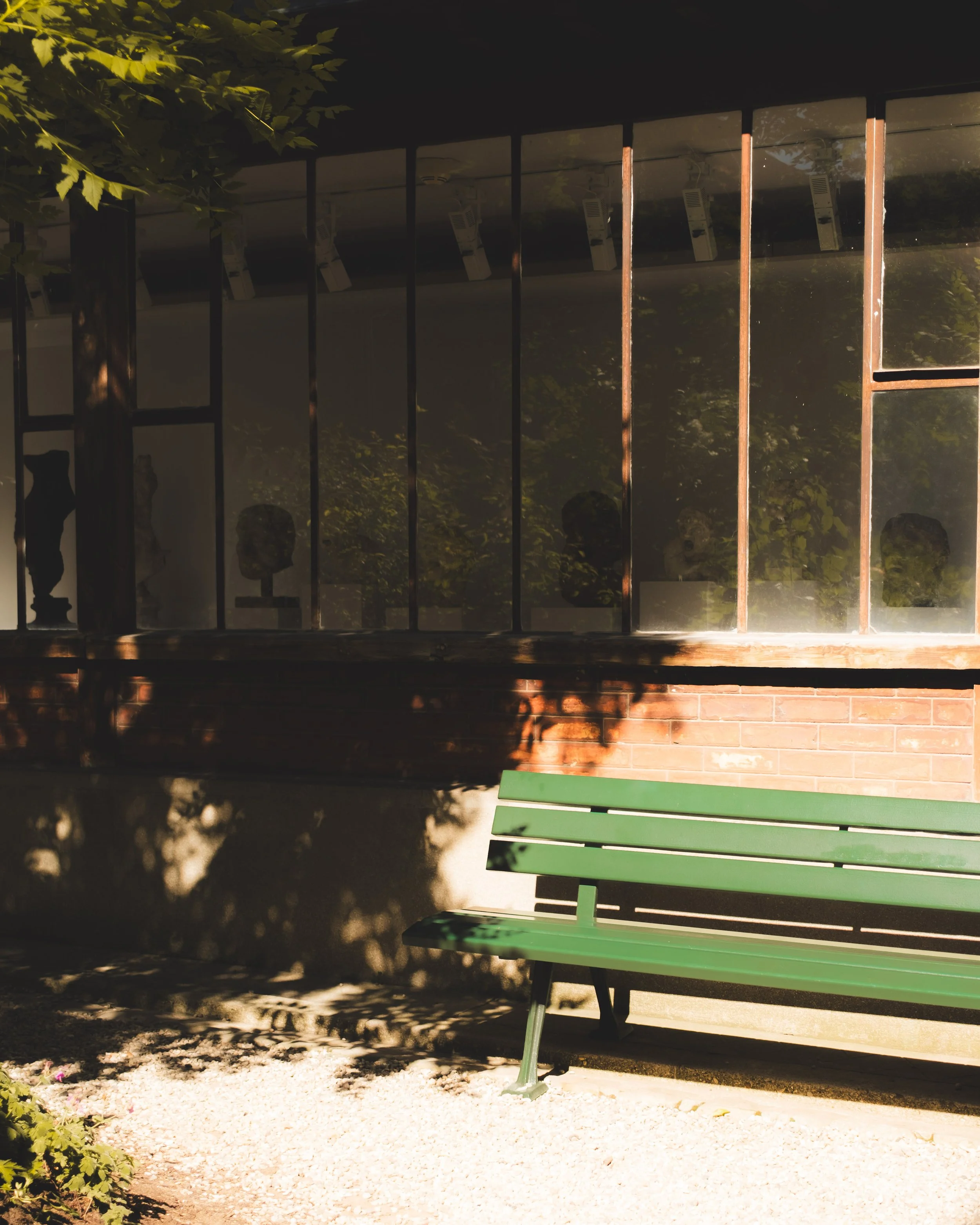 A green park bench with black metal legs is situated on gravel ground near a brick wall with glass windows. The area is shaded with some sunlight filtering through leaves, casting shadows.